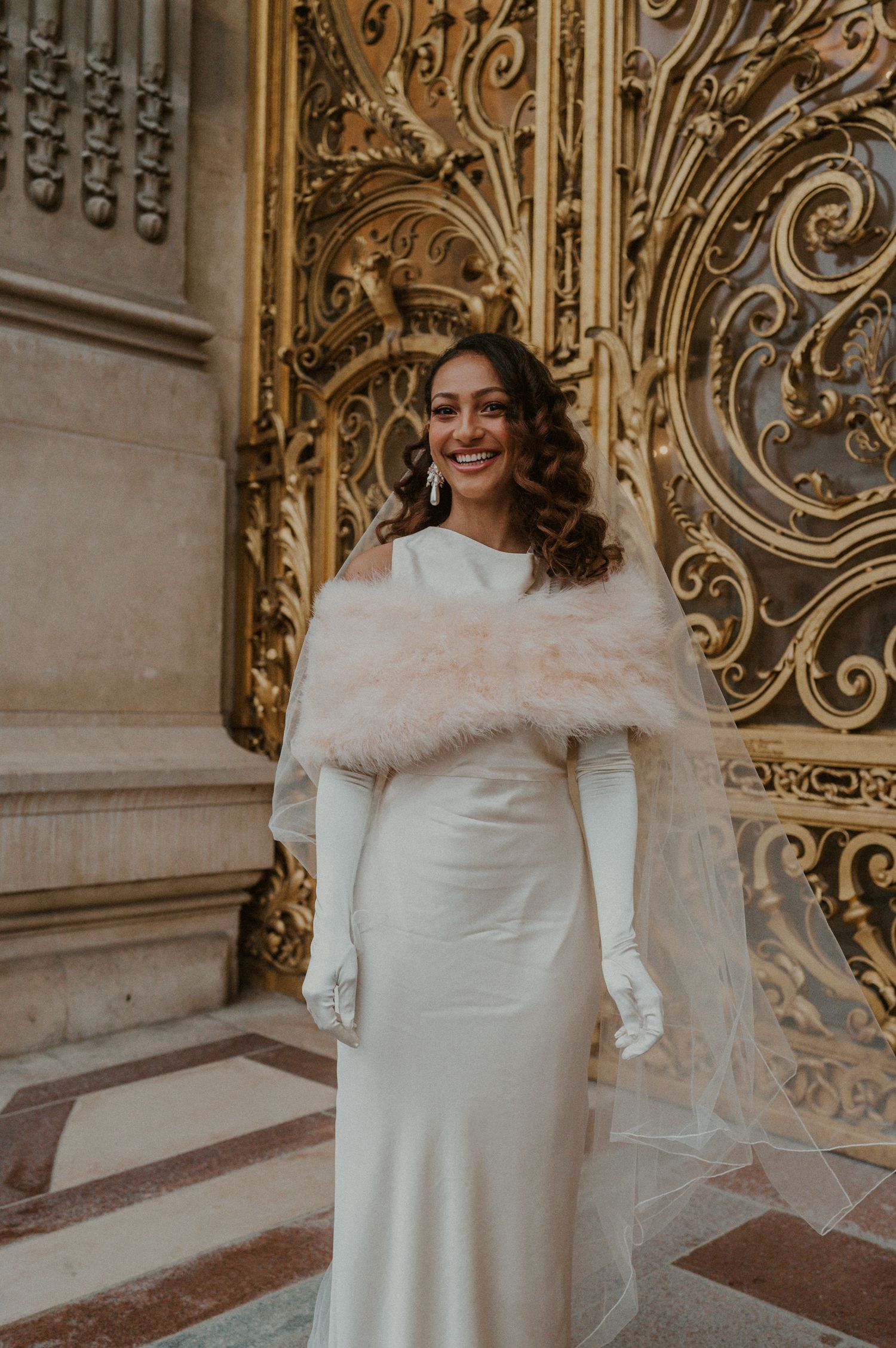 Elegant winter bride wearing a white gown and fur wrap poses against ornate golden gates.