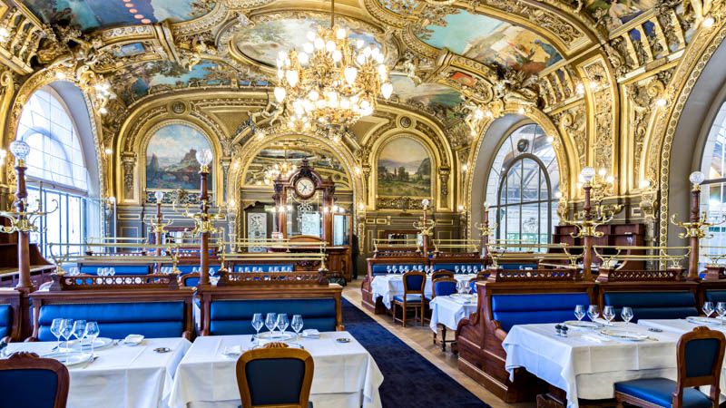 Opulent Belle Epoque restaurant interior with gilded ceiling, chandeliers and white-clothed tables.