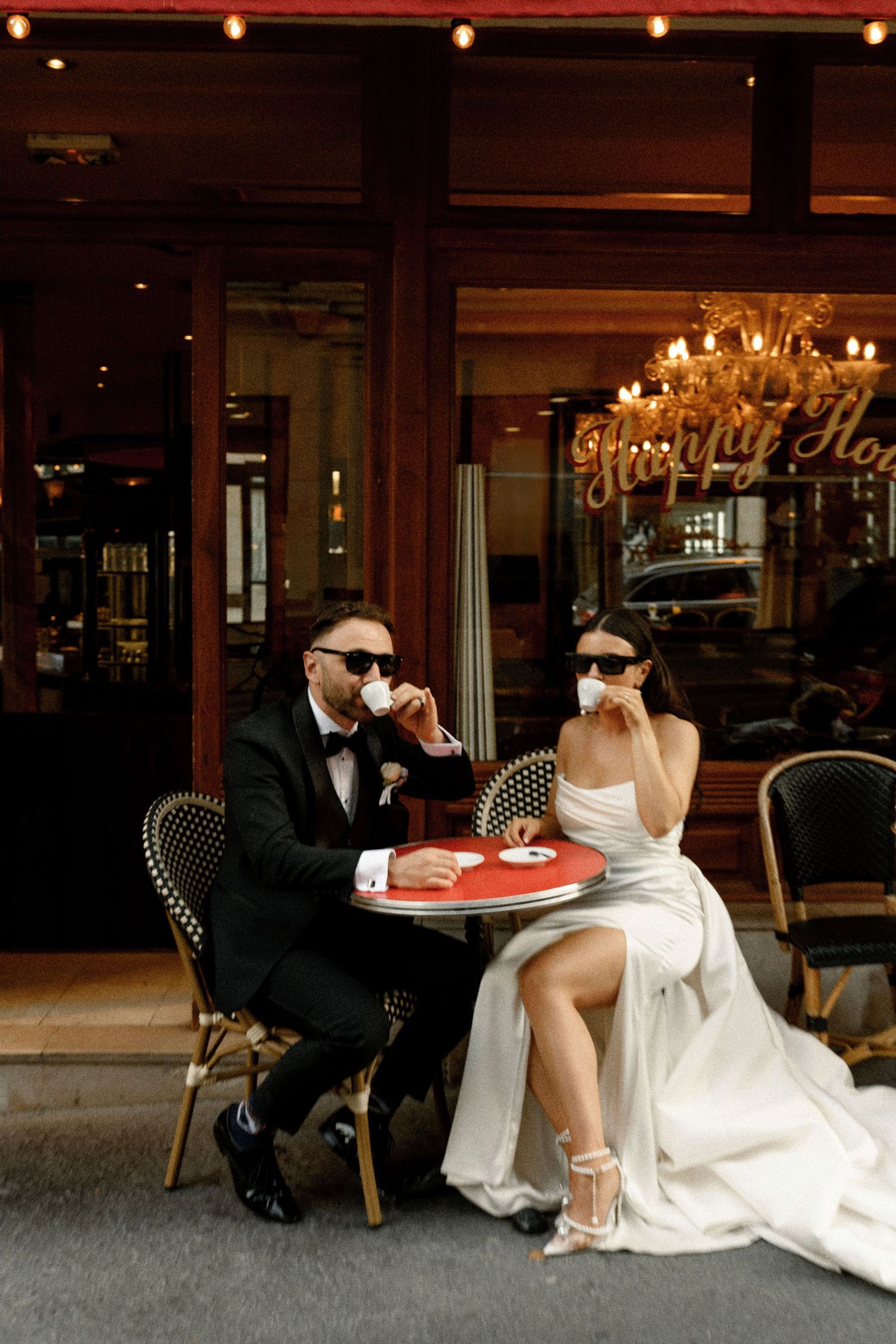 Stylish bride and groom in sunglasses enjoying espresso at a classic Parisian cafe sidewalk terrace.