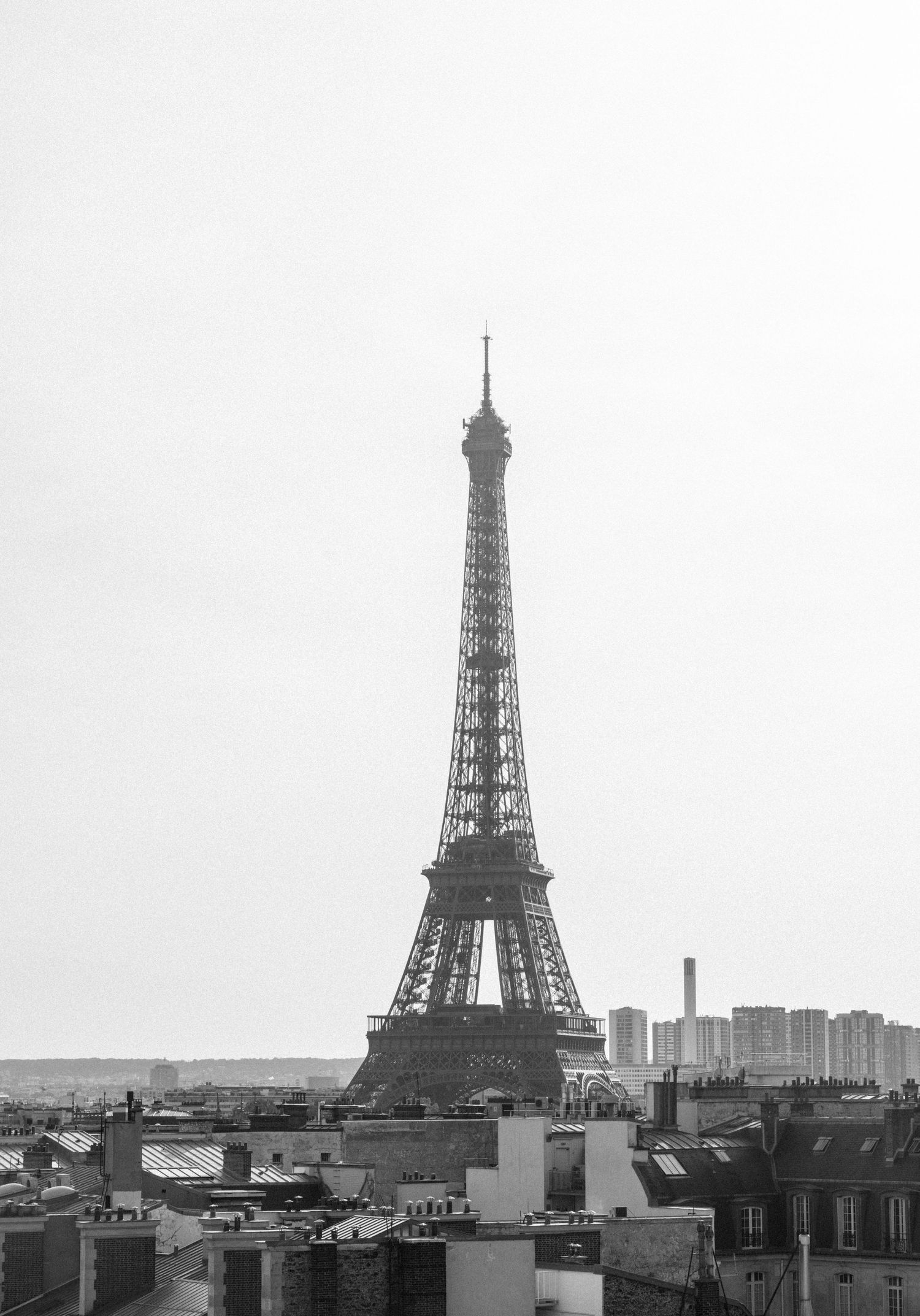 Black and white aerial view of the iconic Eiffel Tower rising above Paris rooftops against a clear sky.