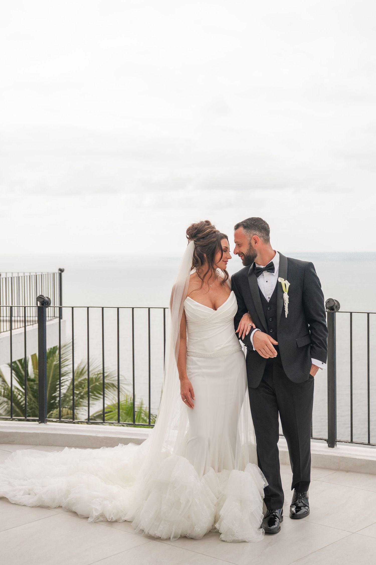 Bride and groom share a romantic moment on a seaside terrace, she in a white gown with veil, he in a black tuxedo.