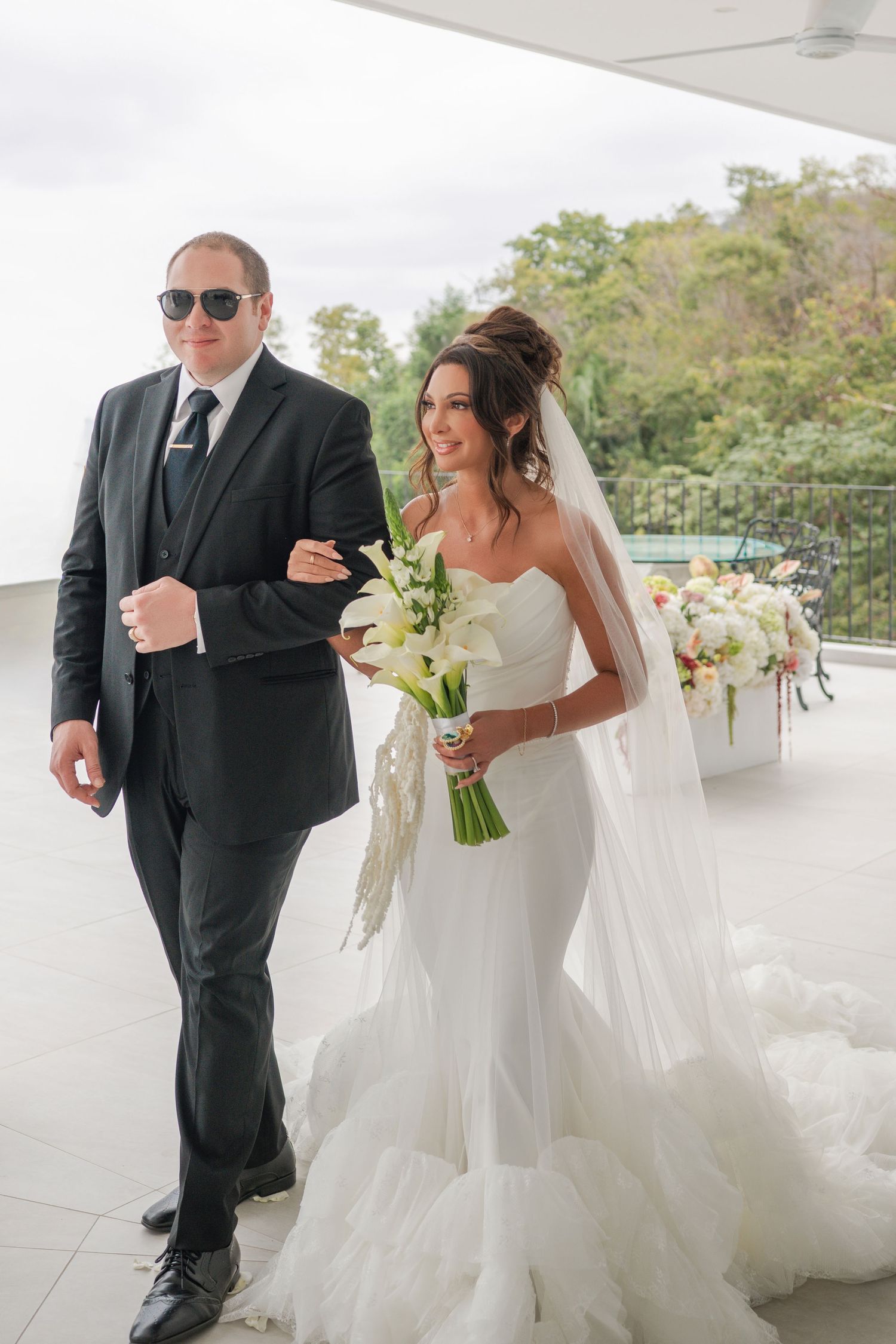 Bride in white strapless gown holding floral bouquet walks arm-in-arm with man in black suit and sunglasses.