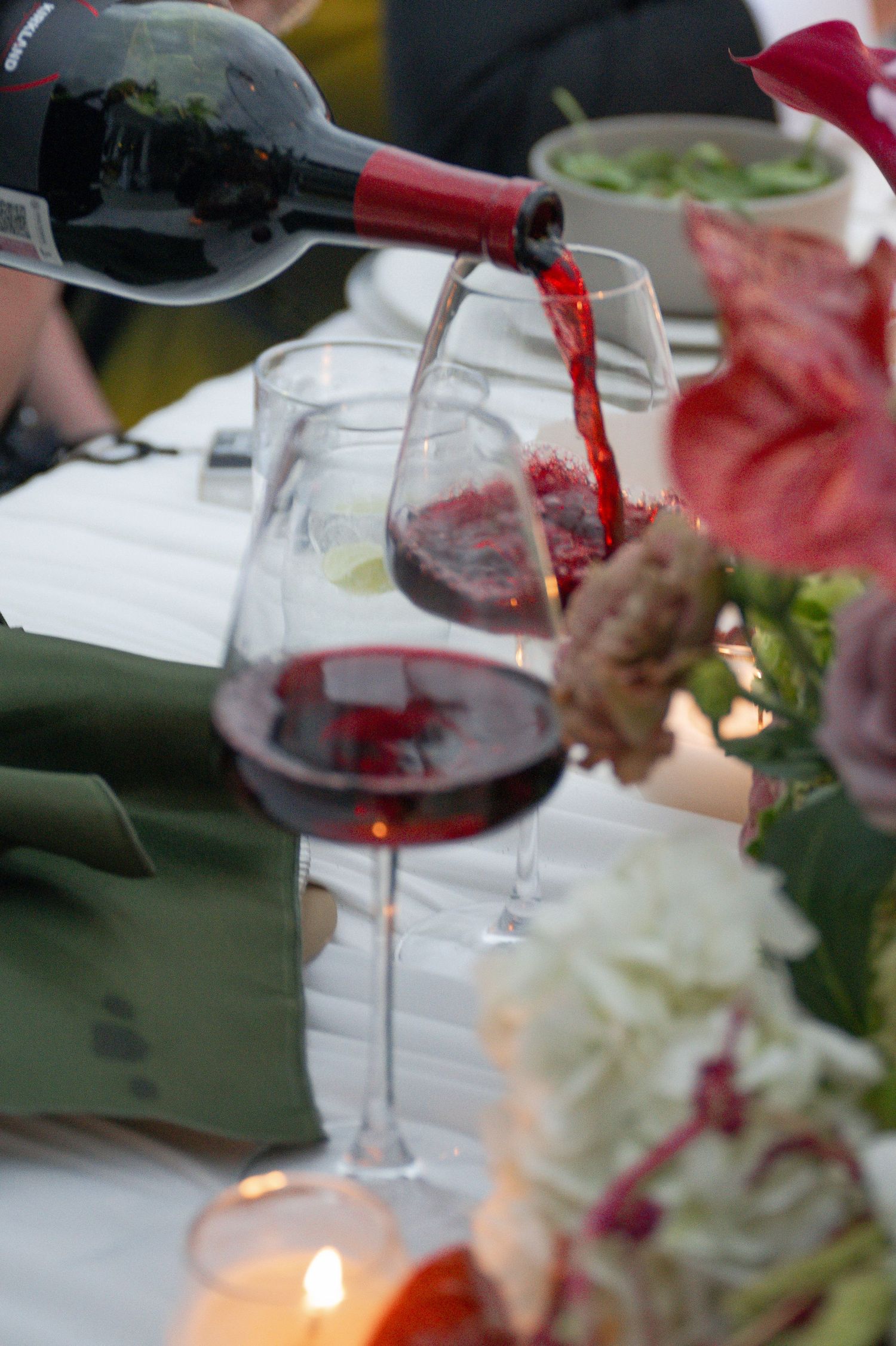 Red wine being poured into a glass at an elegant dinner table with floral arrangements and candles.