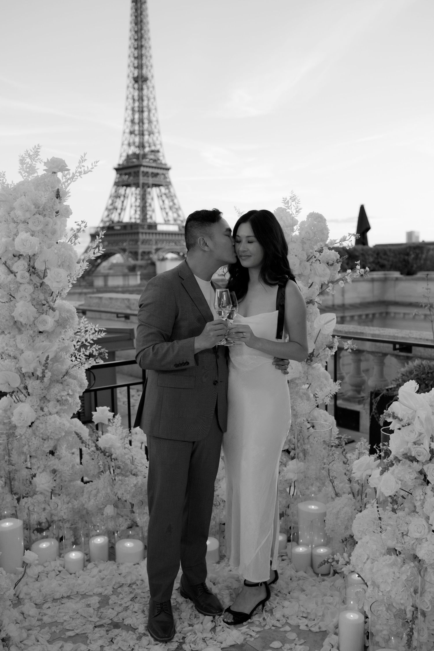 Black and white photo of a couple embracing with champagne on a Paris rooftop, surrounded by white flowers and candles with the Eiffel Tower