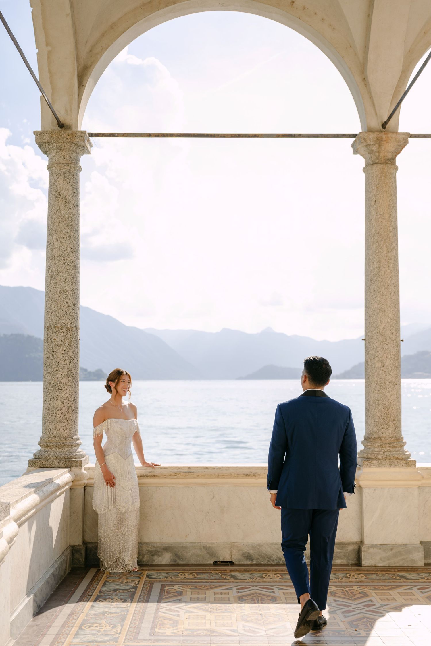 Bride in white gown and groom in navy suit stand under stone arches overlooking a serene lake and mountains.