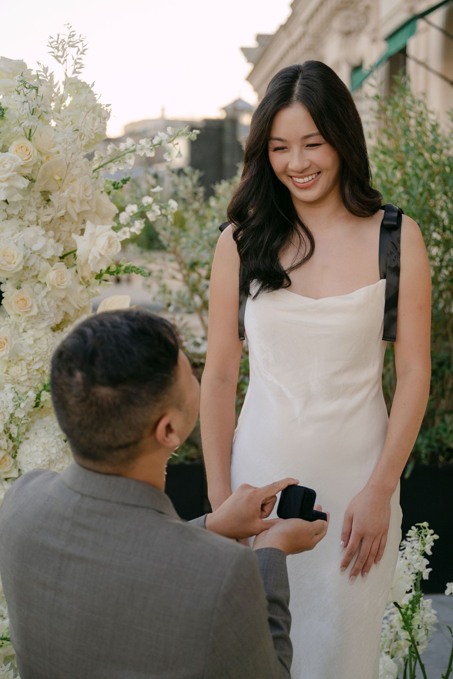Smiling woman in a white slip dress looks down at her partner kneeling with a ring box during a romantic proposal.