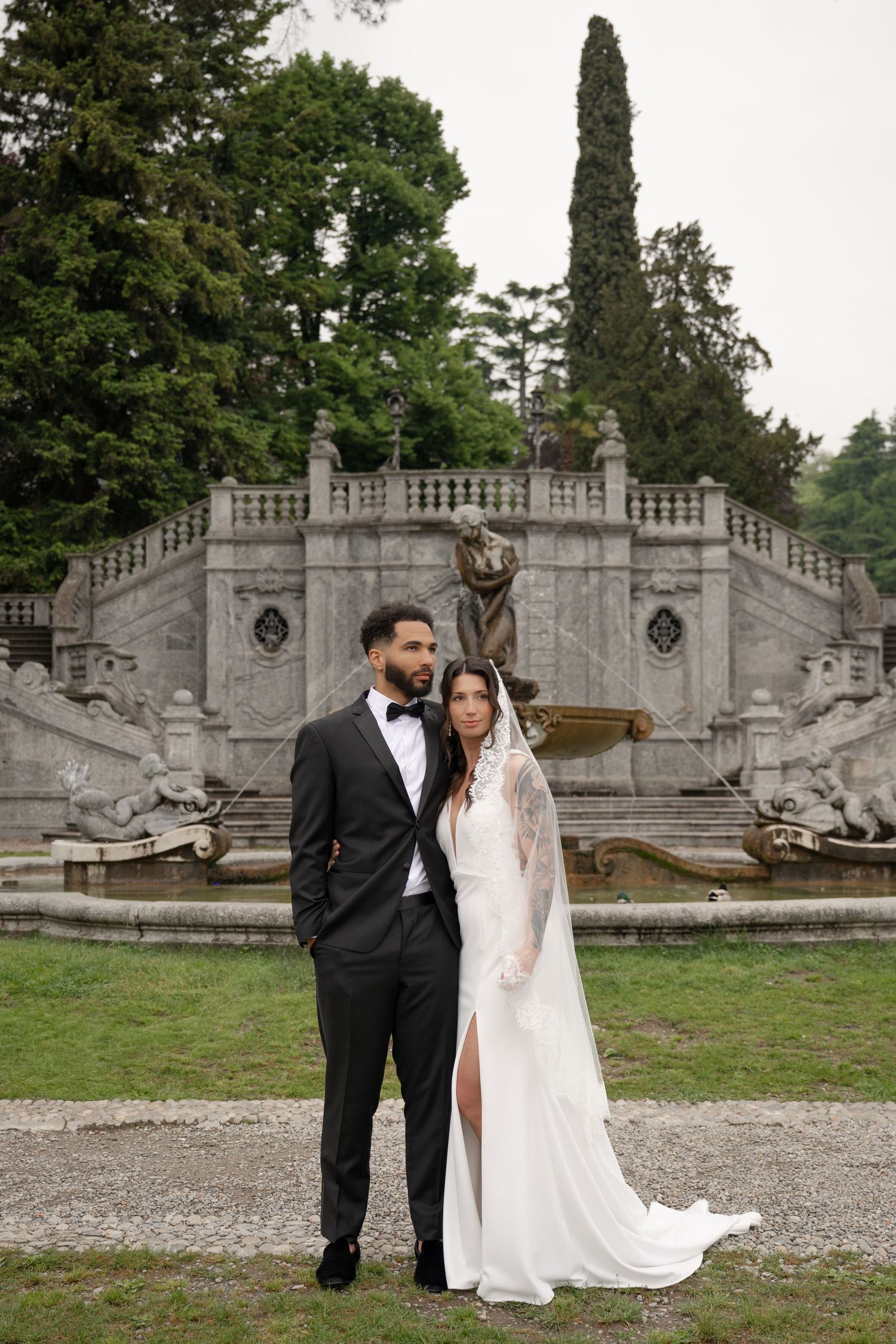 Elegant couple posing before ornate stone fountain in lush garden, bride in lace wedding gown.