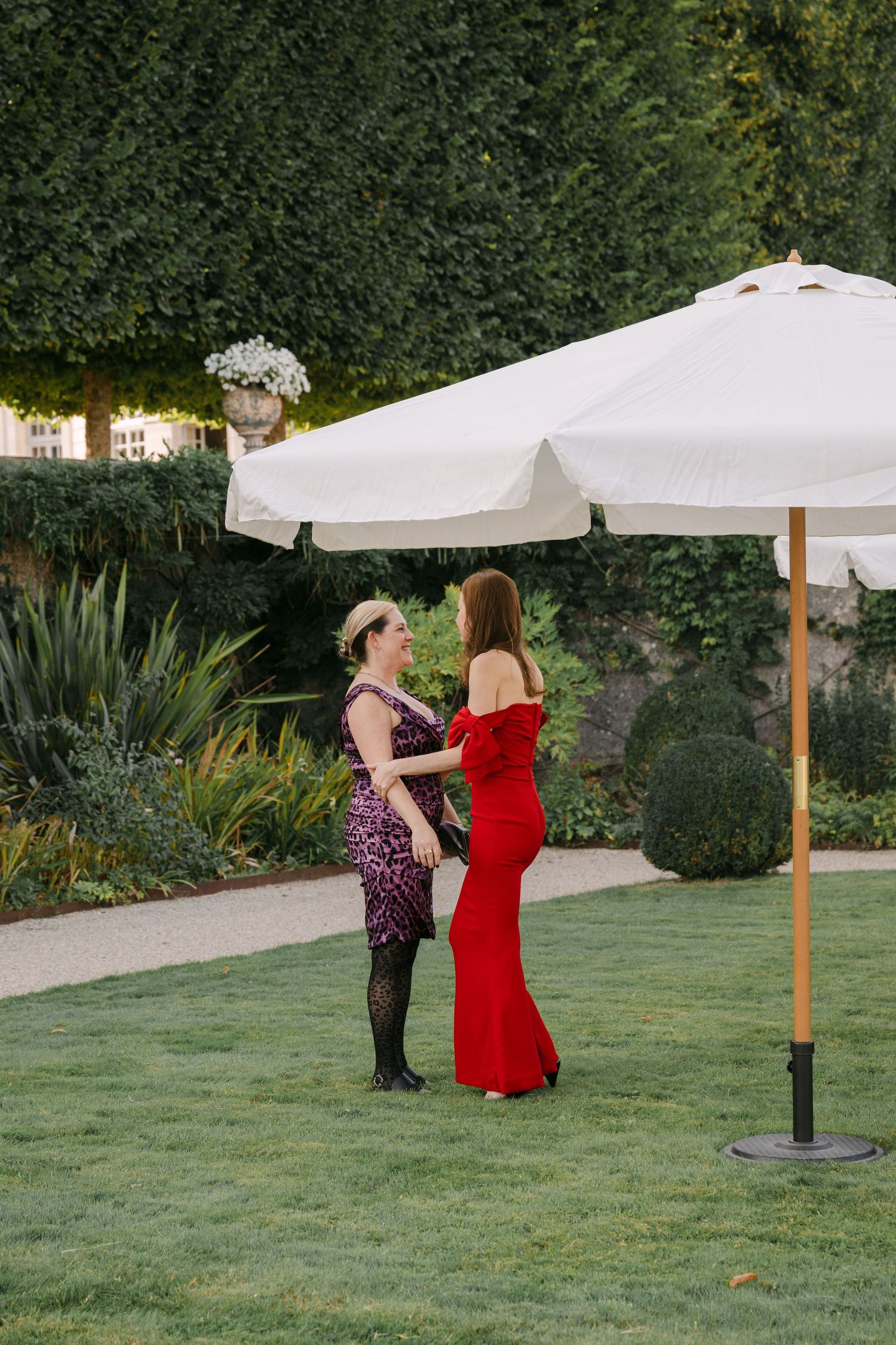 Two elegantly dressed women converse beneath a white market umbrella in a beautifully landscaped formal garden.