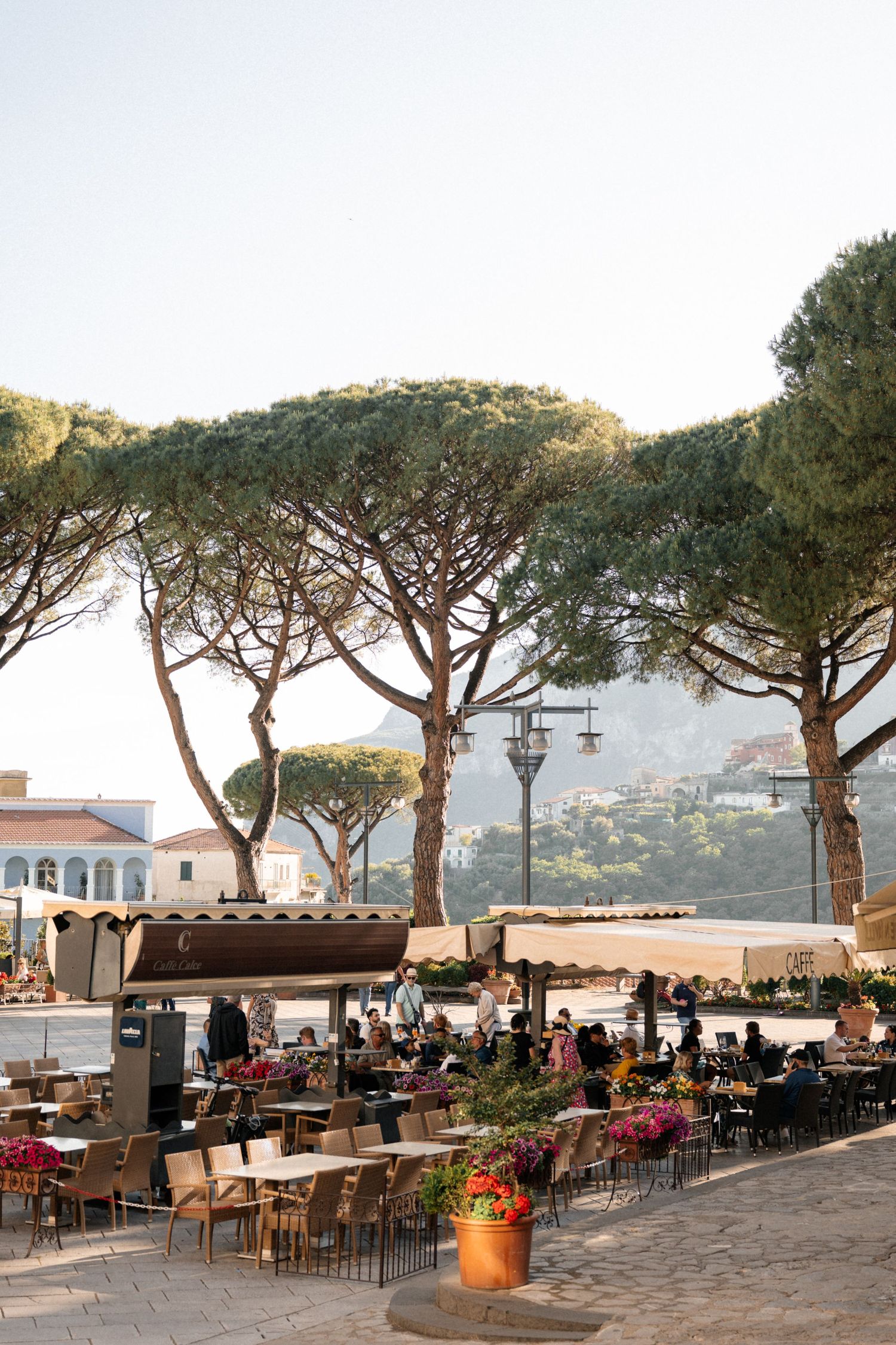 Outdoor Italian cafe terrace with umbrella pine trees, dining guests, and scenic hillside backdrop.