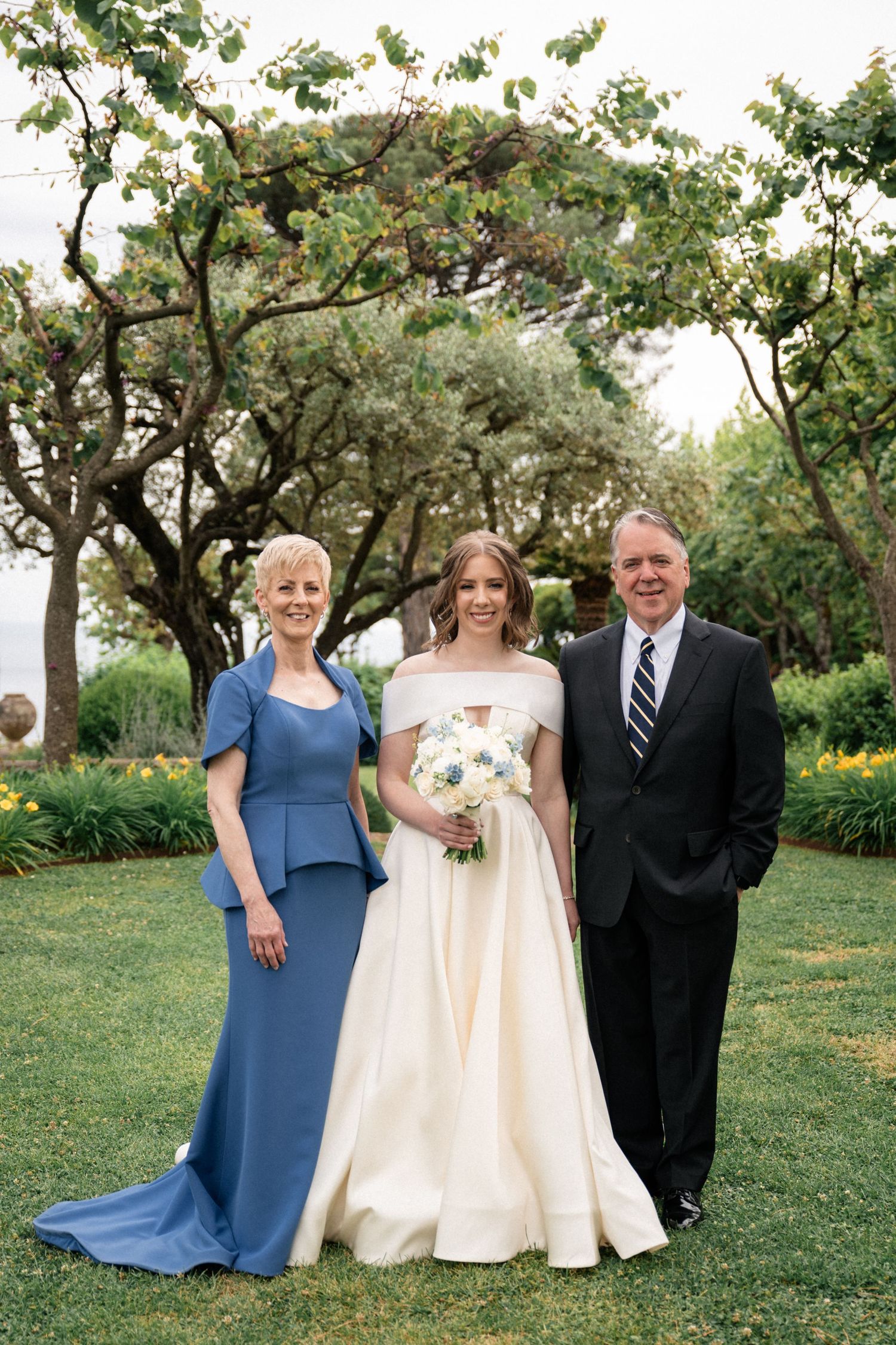 Bride in off-shoulder ball gown holds white bouquet while posing with parents in elegant garden setting.