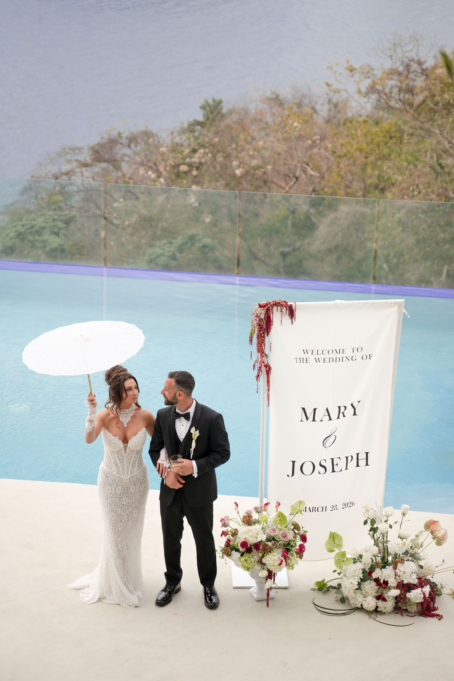 Bride and groom pose by wedding welcome sign for Mary and Joseph near infinity pool with tropical views.