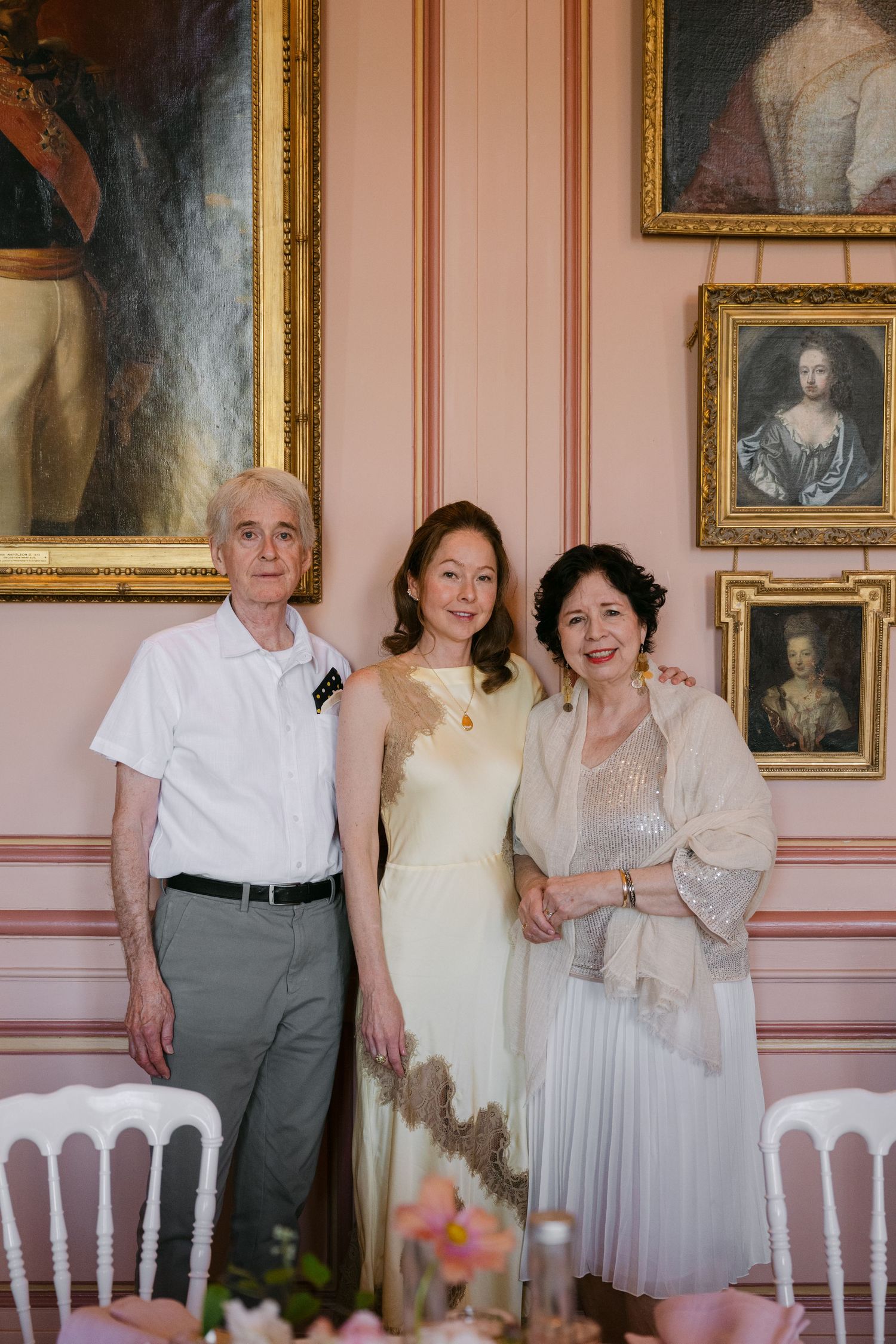 Bride in ivory gown poses with her parents in front of classic oil paintings in a pink-walled French chateau.