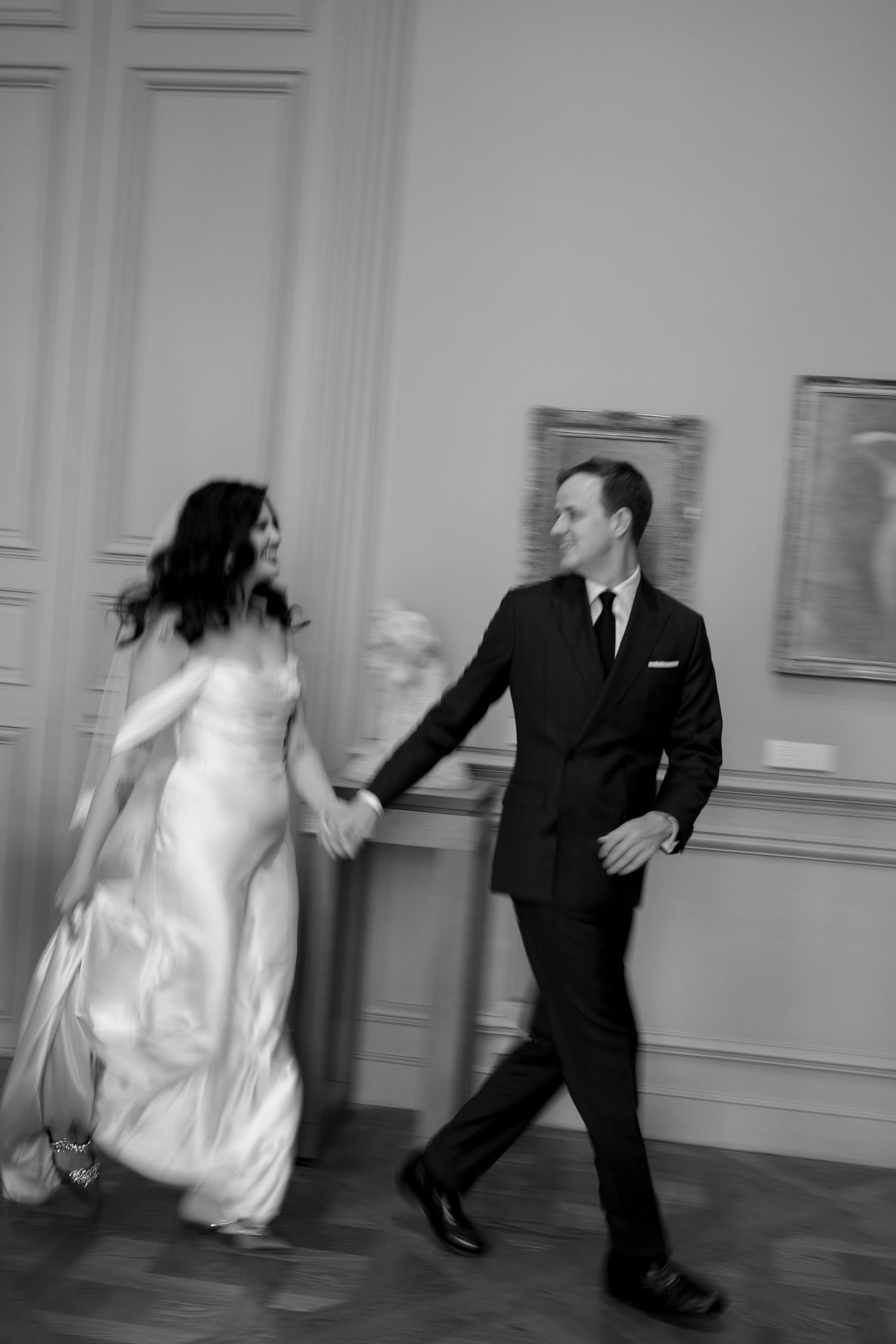 Black and white motion blur photo of a joyful bride and groom holding hands and walking through a classic museum gallery.