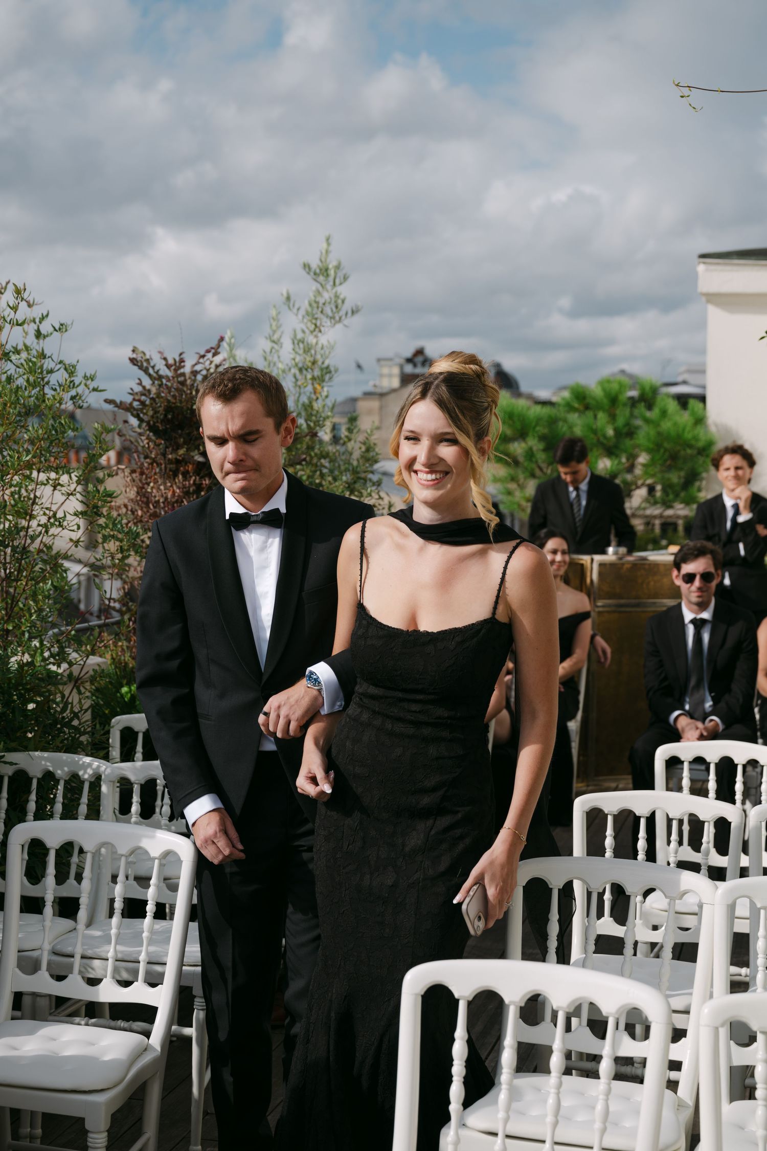 Man in tuxedo escorts elegant woman in black gown down wedding ceremony aisle on Paris rooftop.