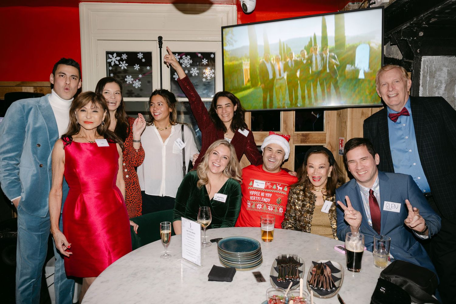Group of eleven people posing together at a holiday party with snowflake decorations and a large screen in background.