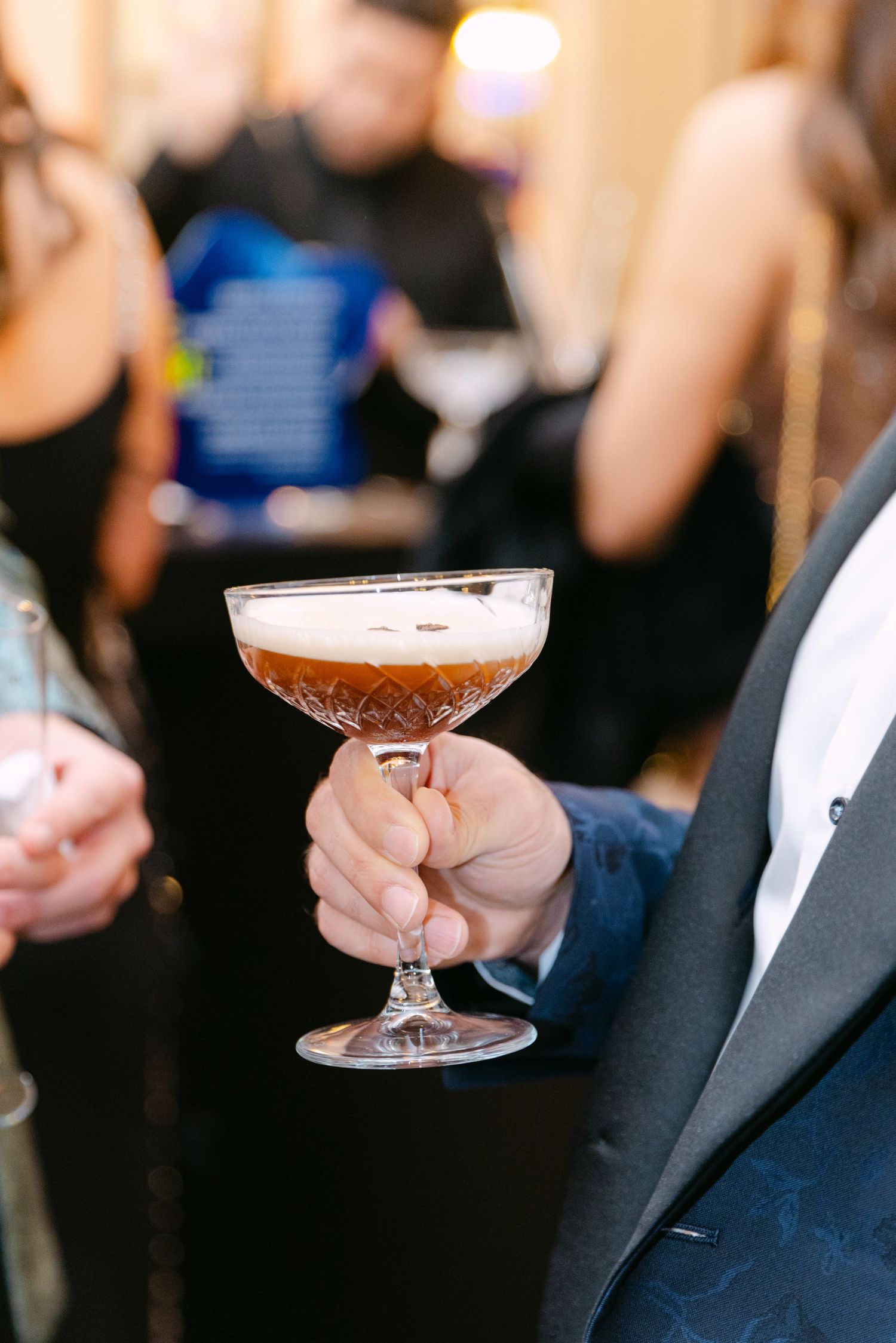 A hand holds a crystal coupe glass filled with a frothy espresso martini cocktail at a formal evening event.