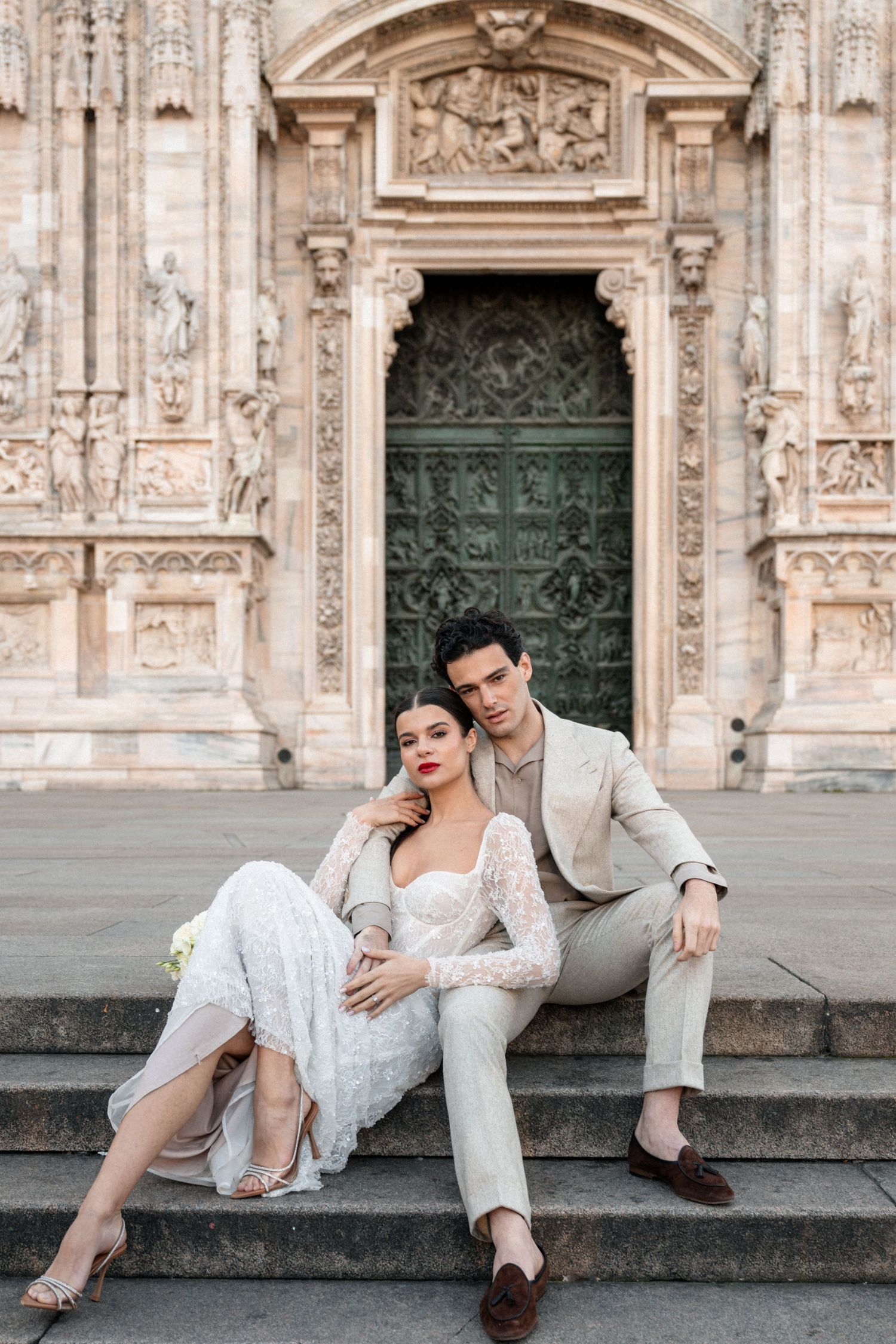 Elegant couple in wedding attire sitting on steps before the ornate doors of Milan Cathedral.