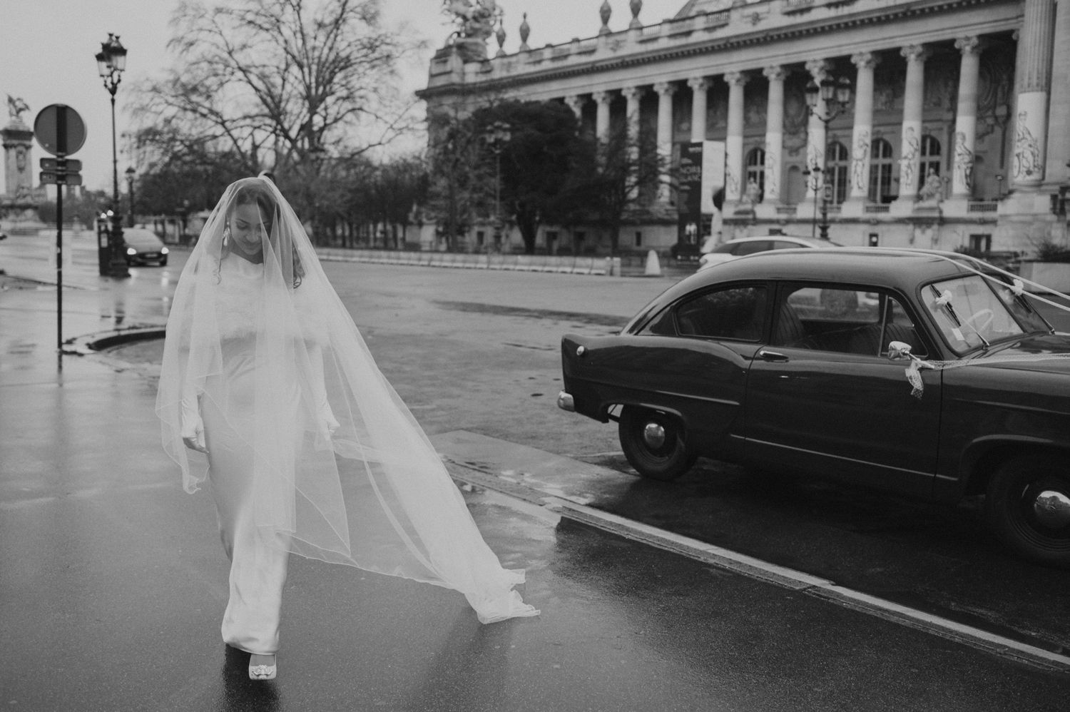 A vintage black and white photo of a bride walking past a classic car in the rain.