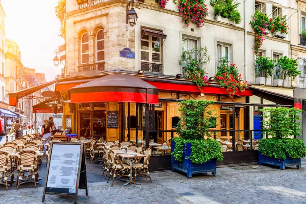 Charming Parisian cafe with red awning and outdoor seating on a cobblestone street corner at sunset.