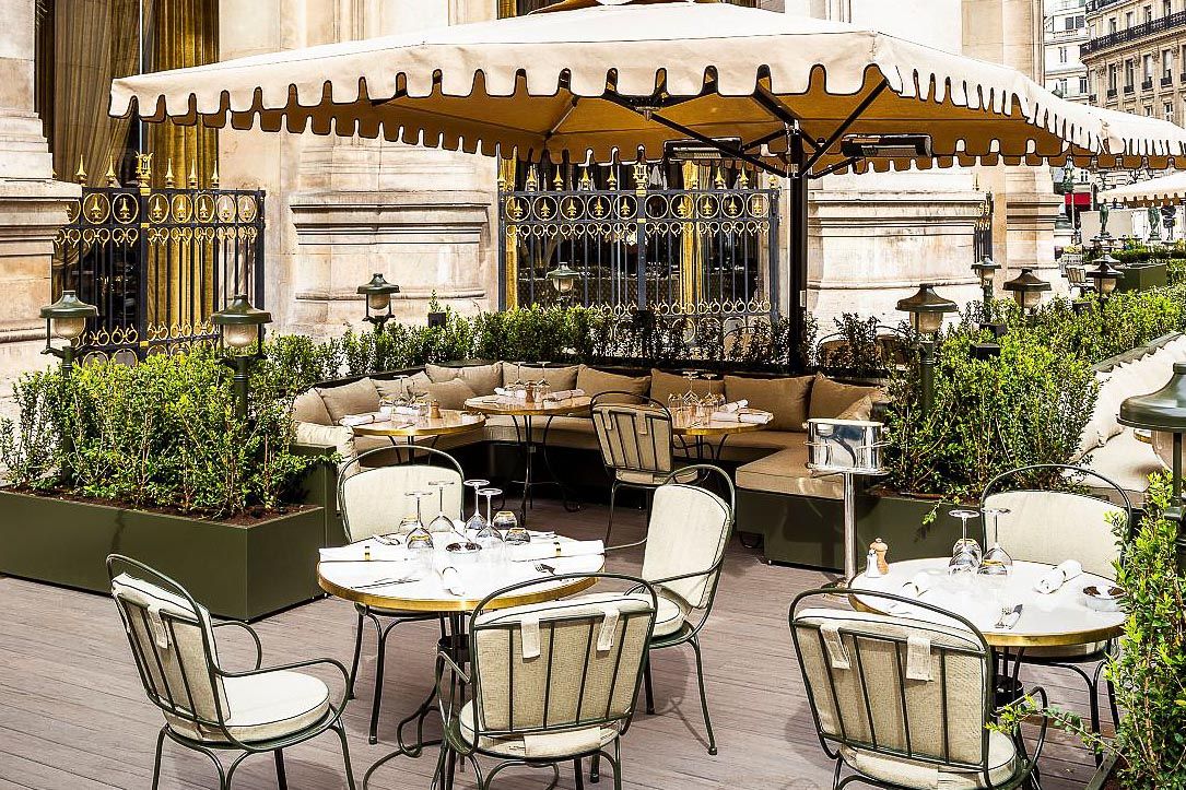 Elegant outdoor cafe seating with metal chairs and planters under cream-colored awning.