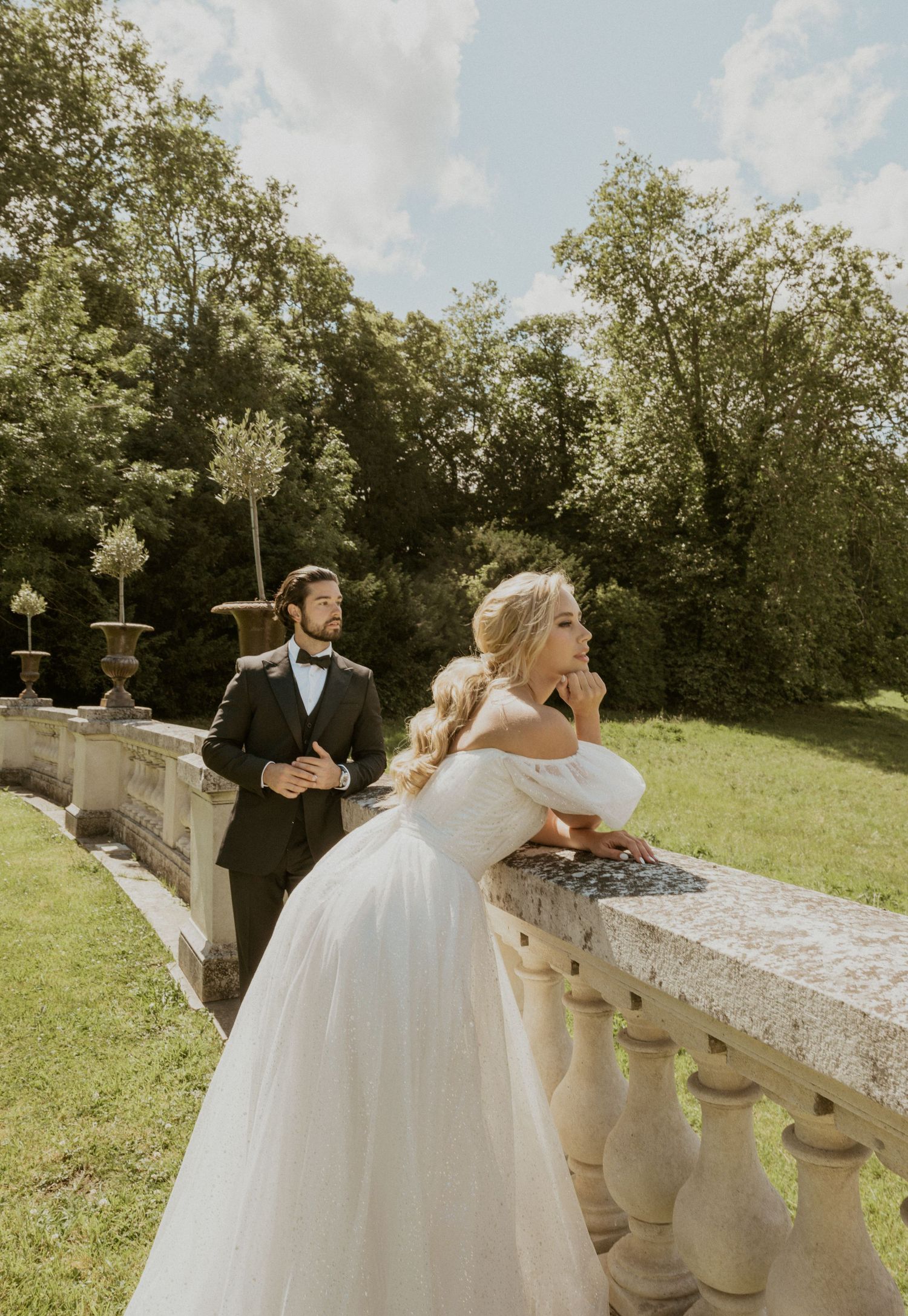 A couple shares a tender first look moment on an elegant stone balcony in a garden setting.