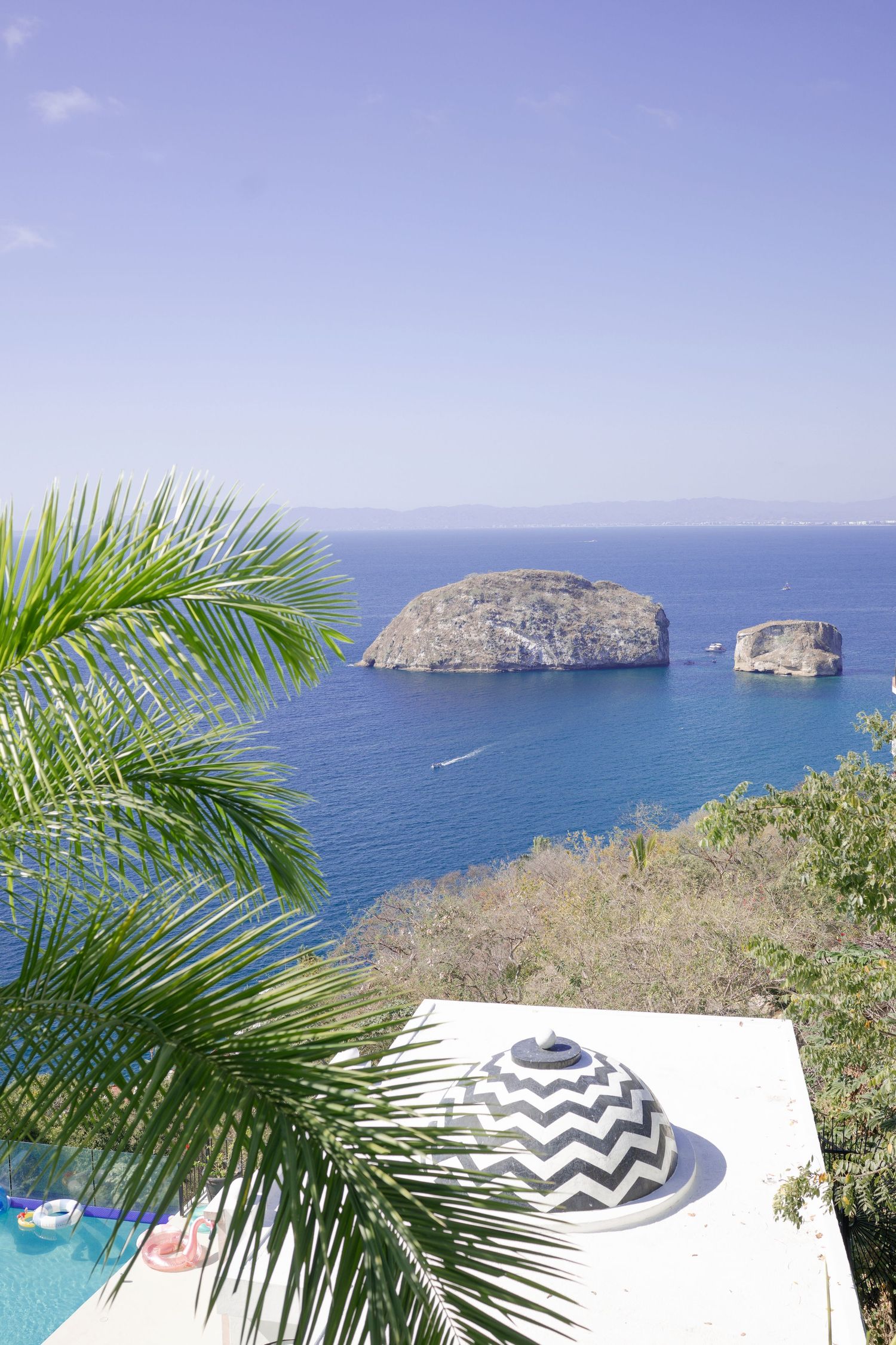 Tropical ocean view with rocky islands, palm fronds, and a chevron-patterned hat on a white ledge under clear blue sky.