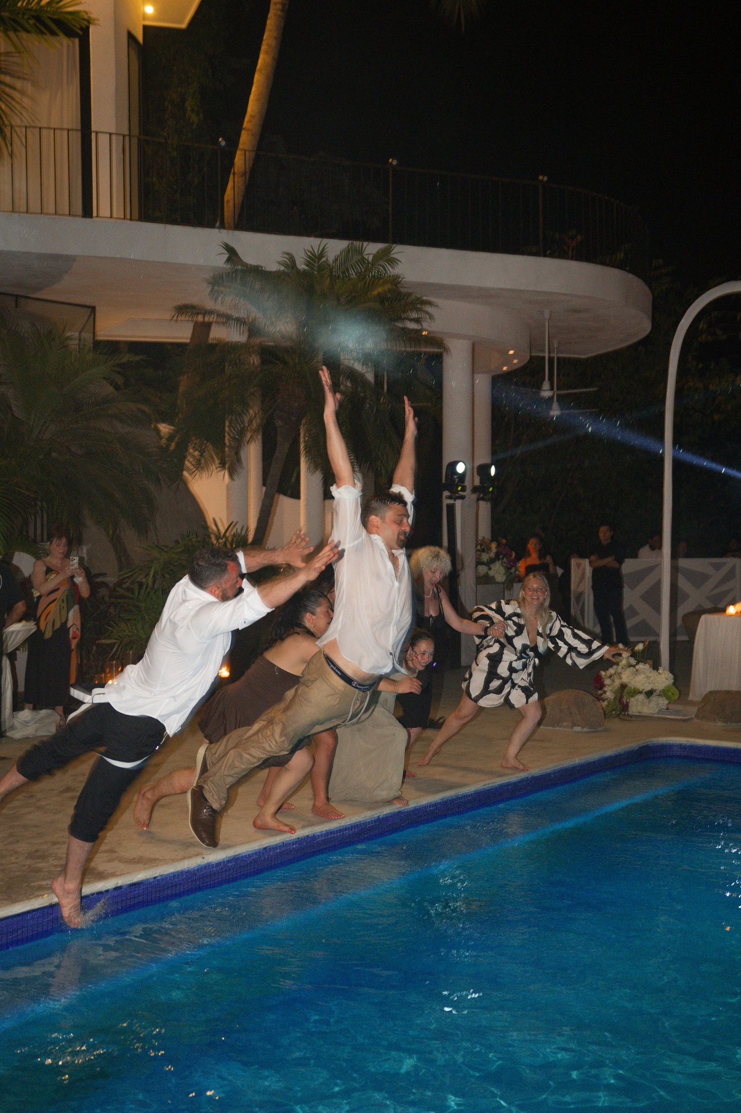 Group of people jumping into a blue swimming pool at night during an outdoor party near a tropical resort.