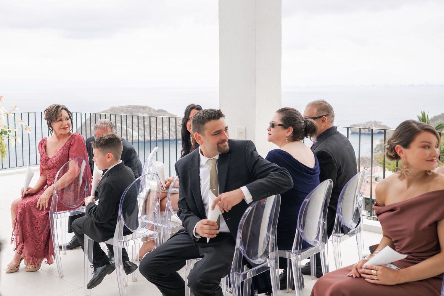 Guests seated in clear chairs at an outdoor wedding ceremony on a terrace with ocean views.