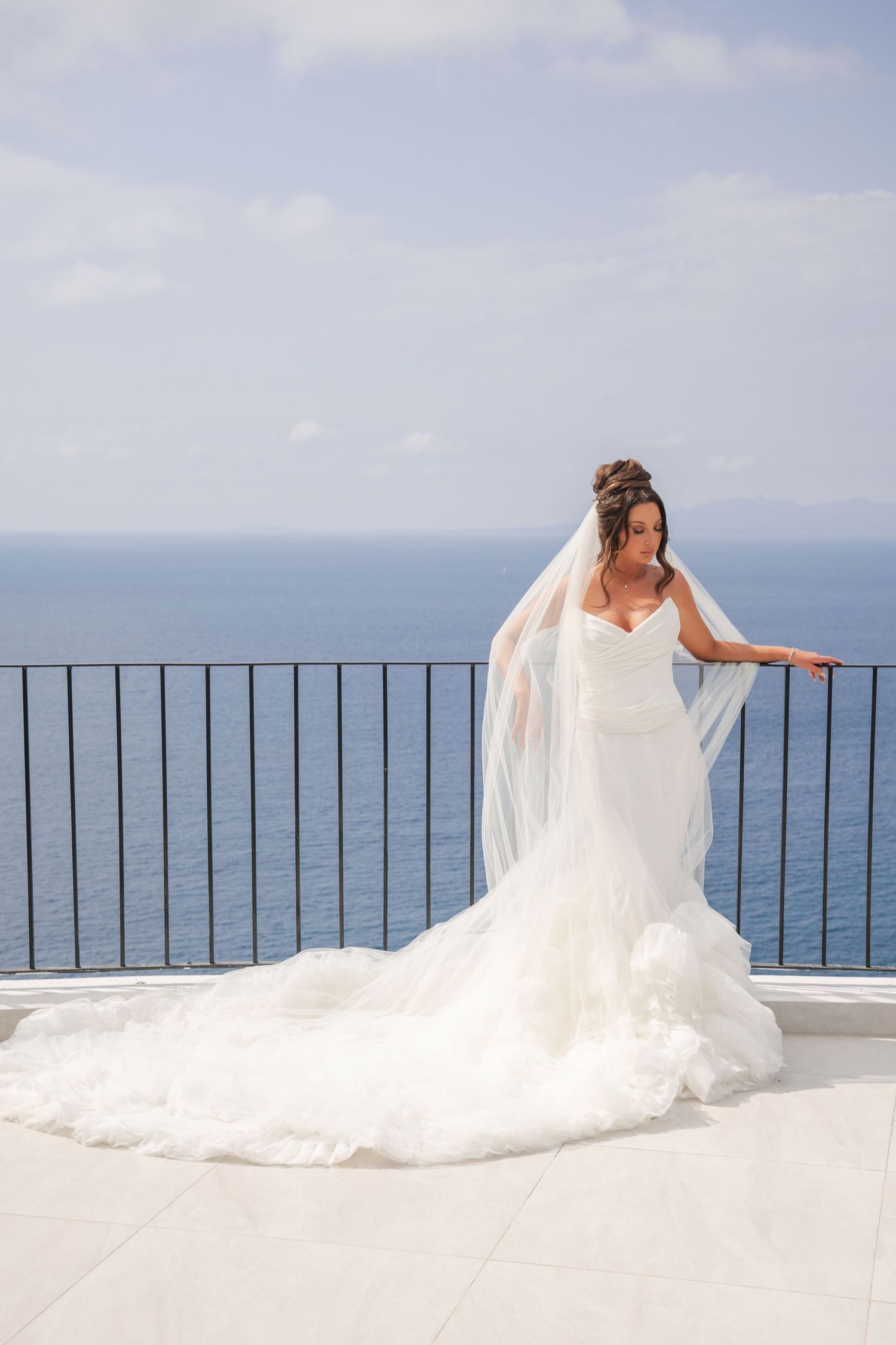 Bride in a white mermaid gown and veil poses on a seaside balcony with ocean views.
