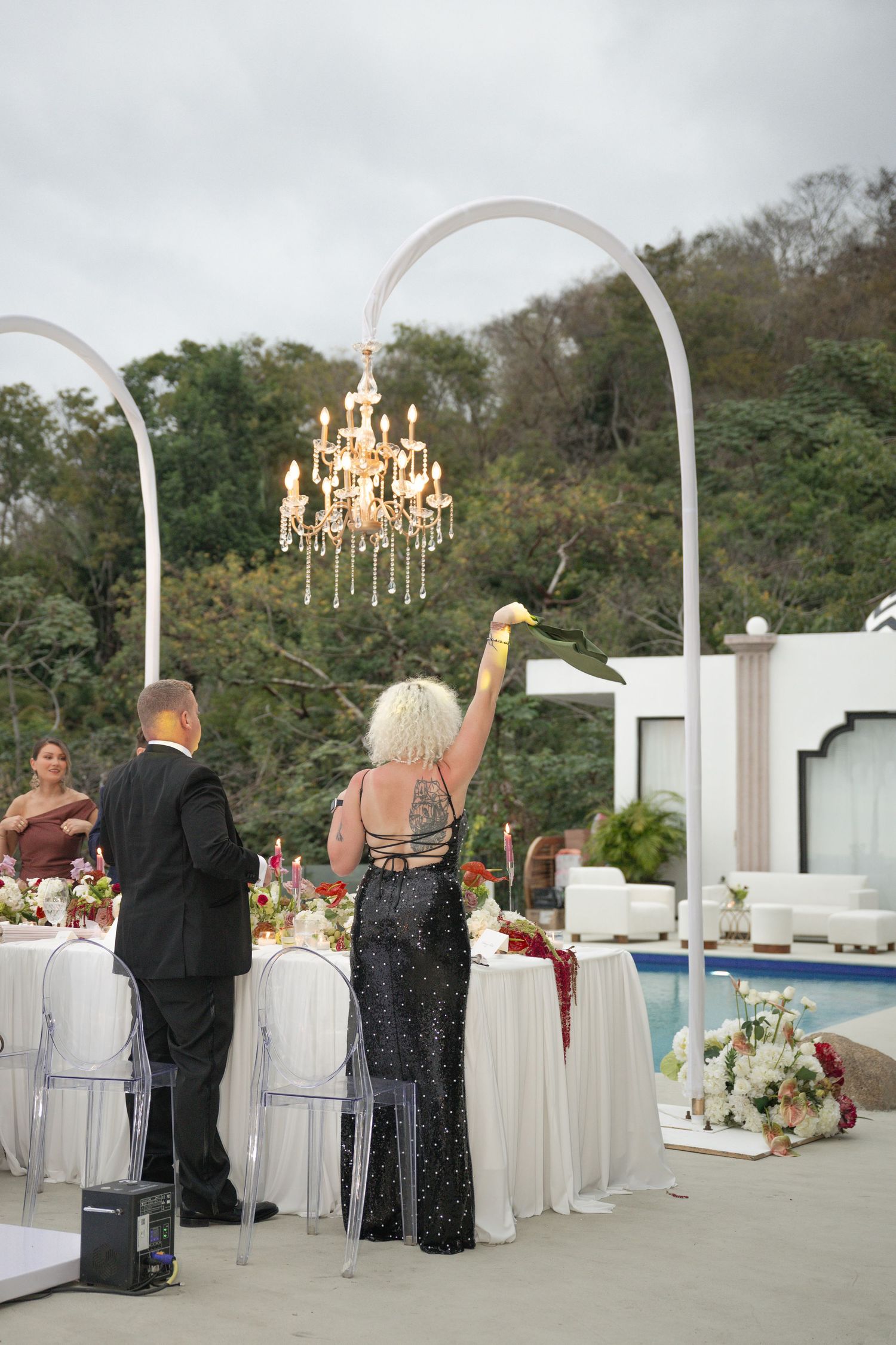 Elegant outdoor wedding reception with chandelier arch, guests in formal attire, pool, and lush greenery backdrop.