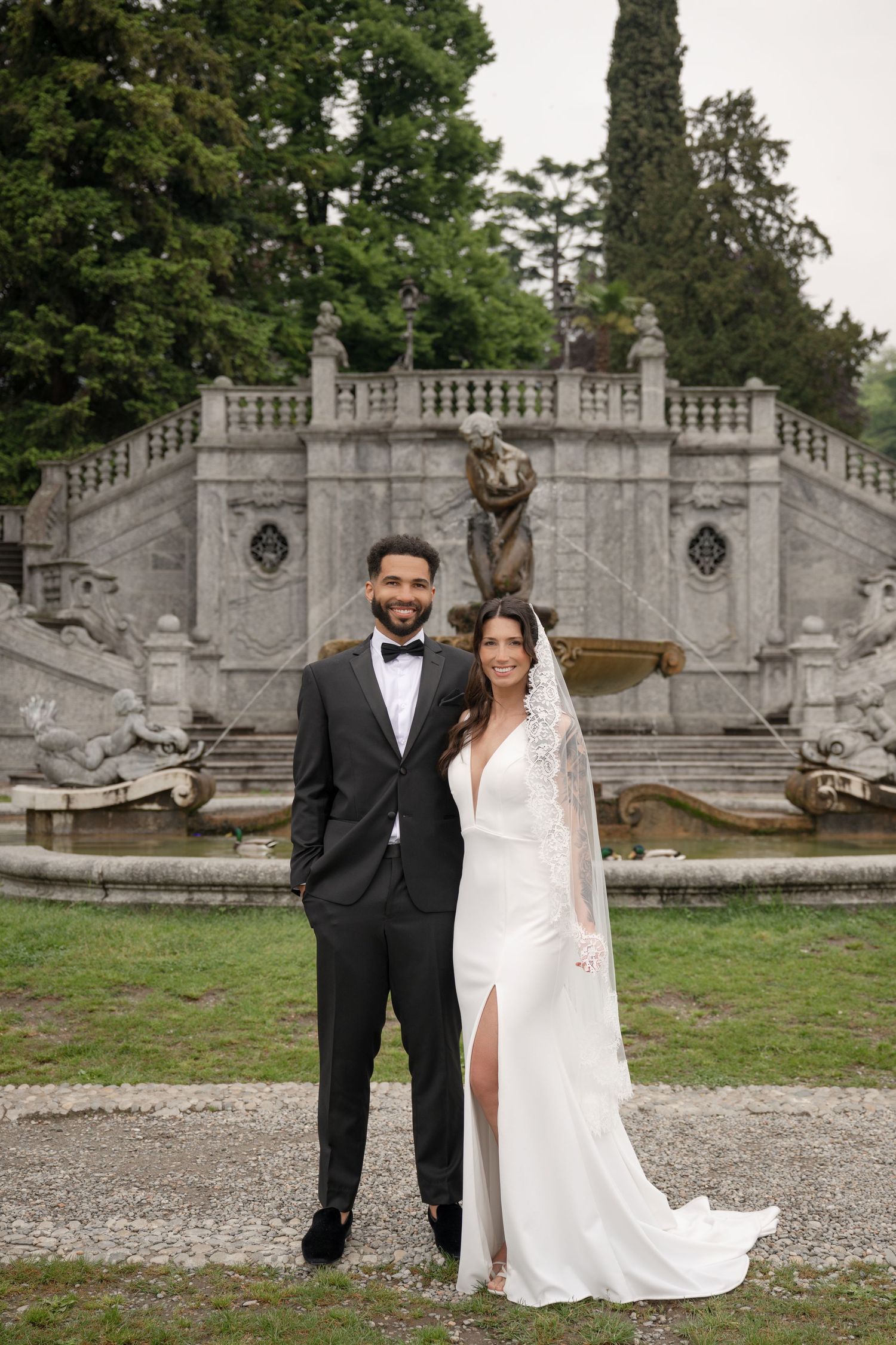 Bride and groom pose together in front of ornate stone fountain in lush garden setting.