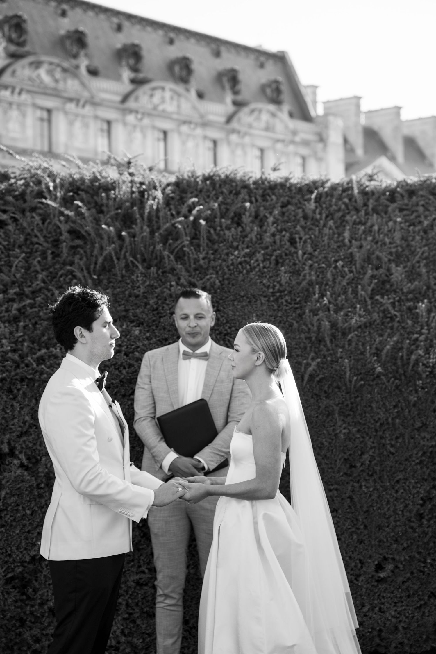 Bride and groom hold hands at outdoor ceremony before green hedges and historic building.