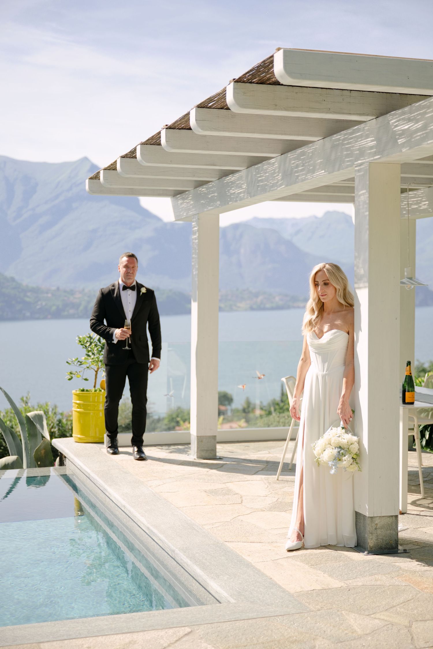 Elegant bride in white gown and groom in tuxedo by a pool with stunning mountain lake views at an outdoor wedding venue.