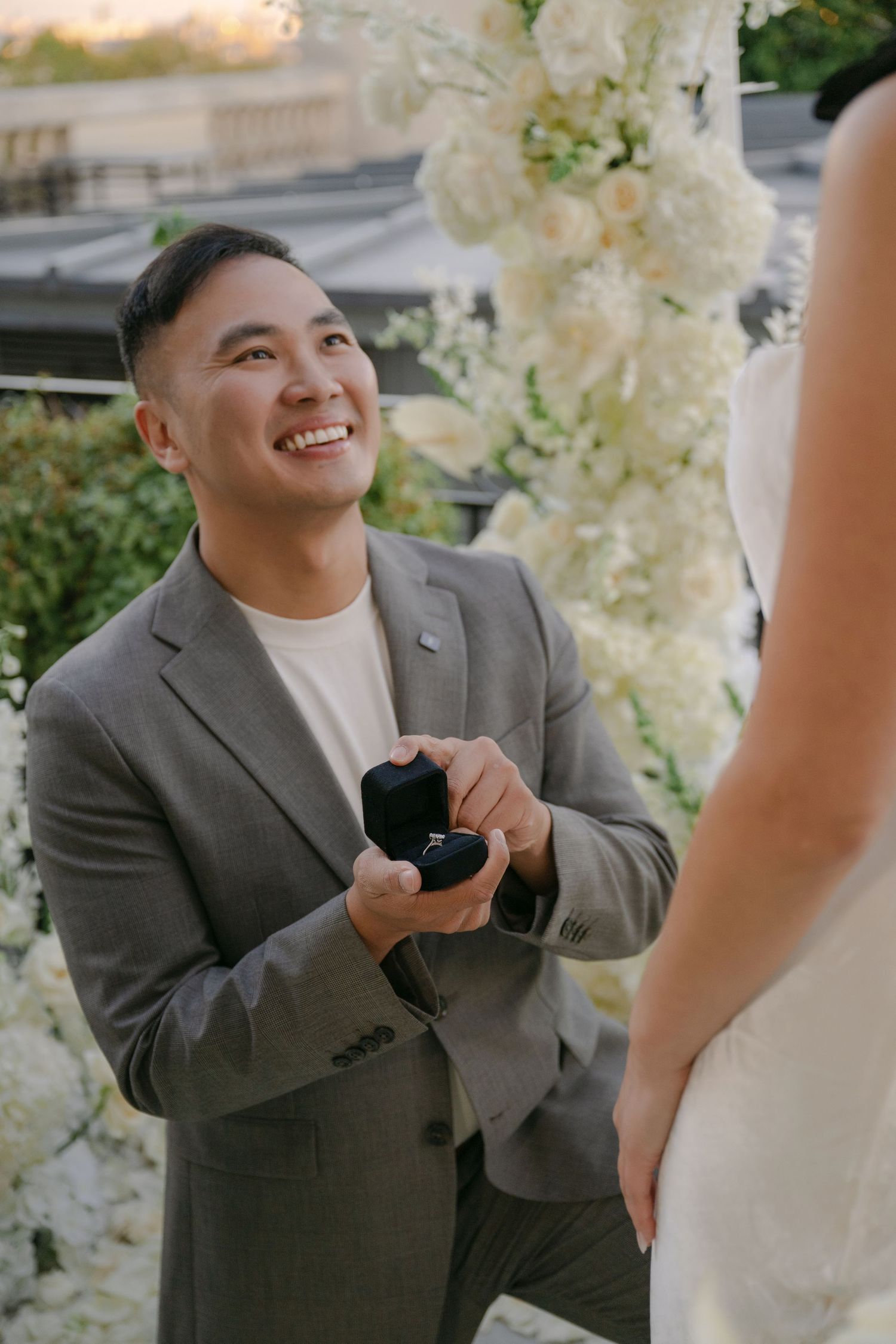 Smiling man in a gray blazer kneels holding a ring box during a surprise proposal surrounded by white flowers.