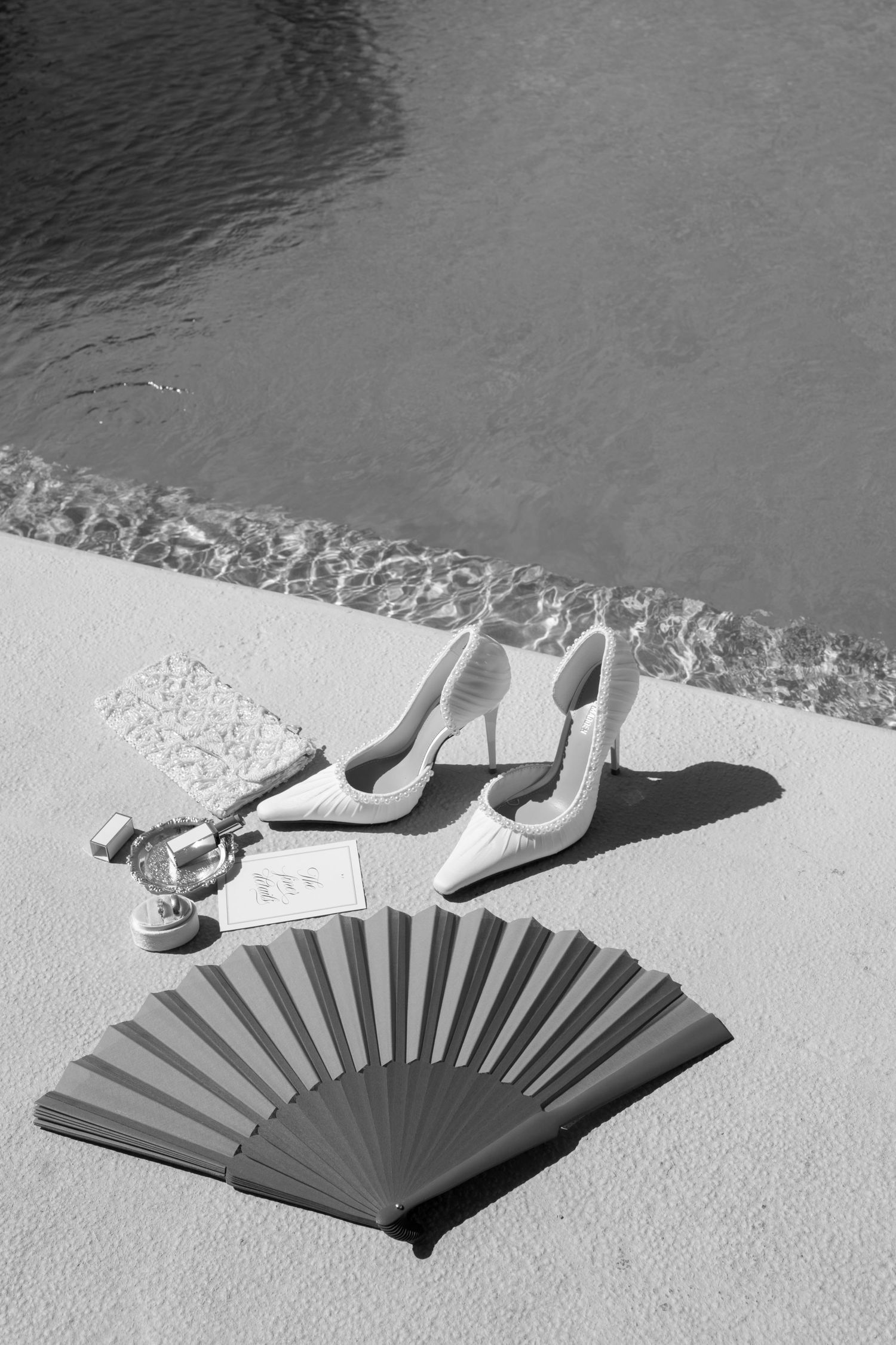 Black and white photo of bridal accessories including heels, fan, and jewelry arranged by a poolside.