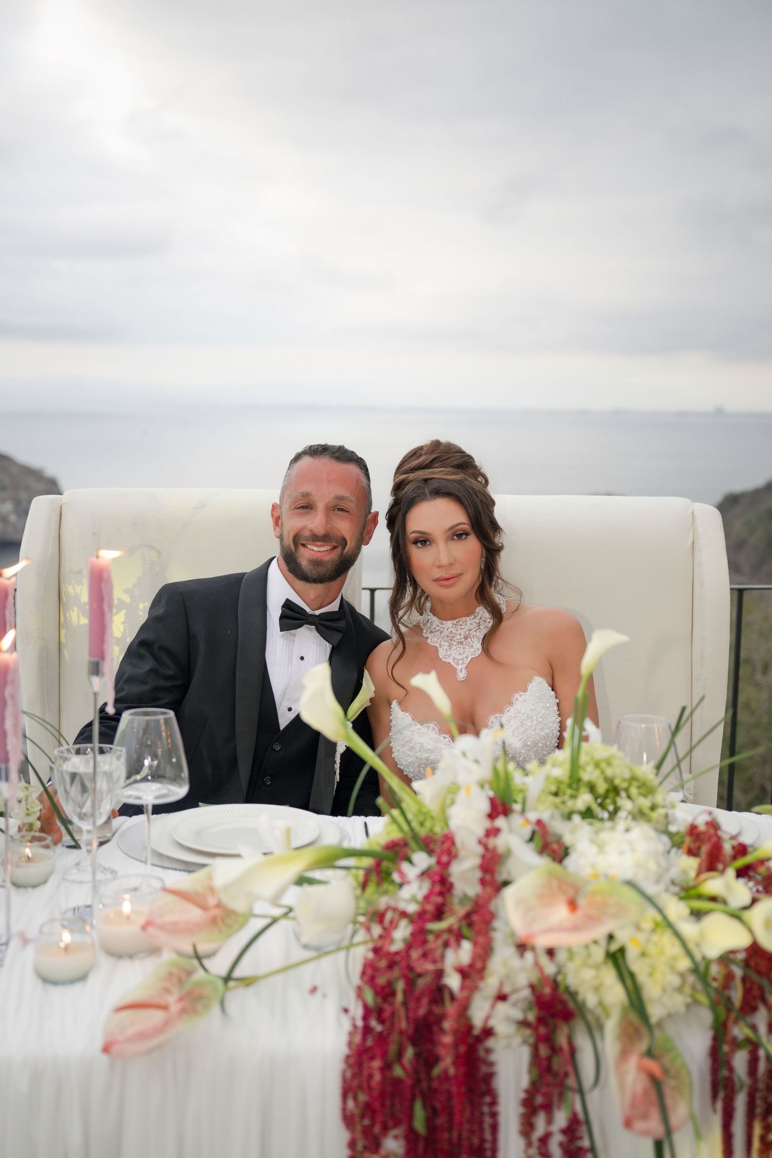 Smiling bride and groom seated at sweetheart table with floral centerpieces and ocean backdrop.