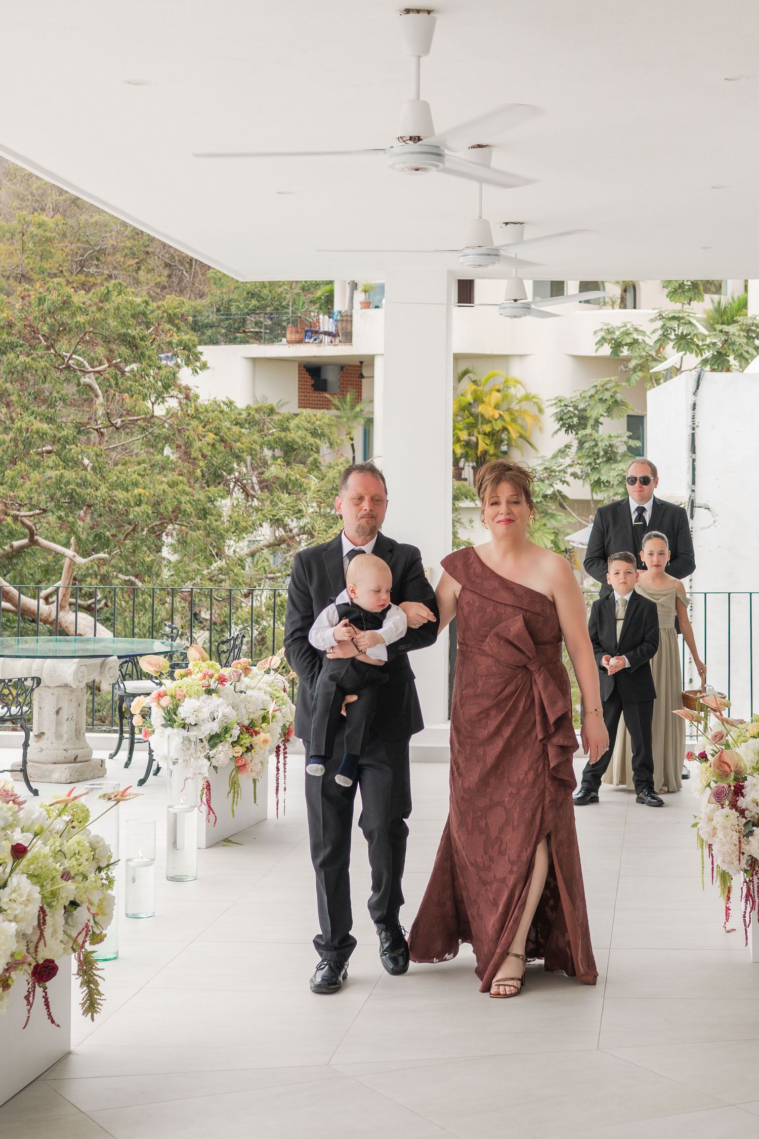 A man in a tuxedo carrying a baby walks alongside a woman in a brown gown down a floral-lined wedding aisle.