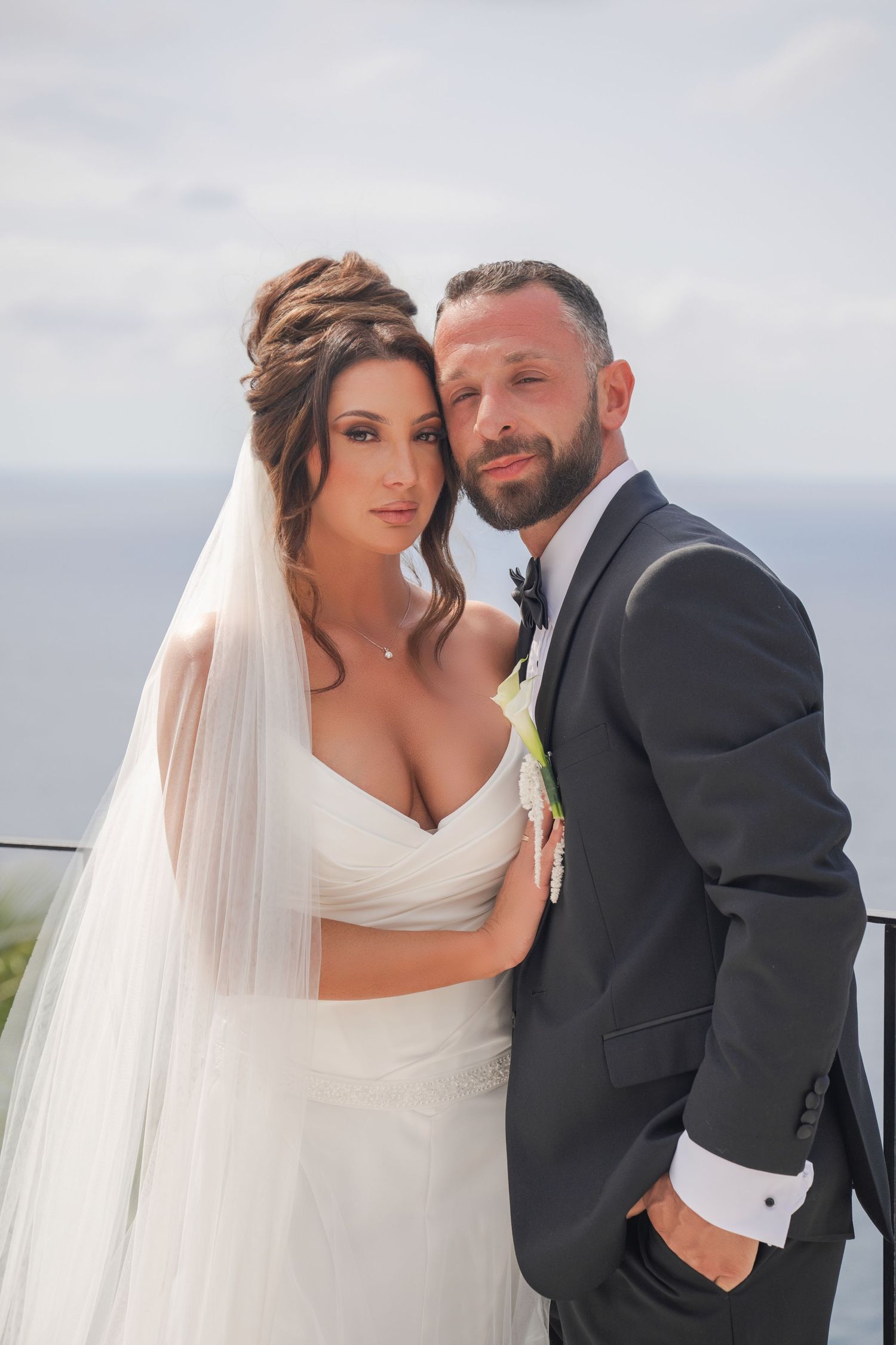 Bride and groom pose together on a balcony overlooking the ocean on their wedding day.