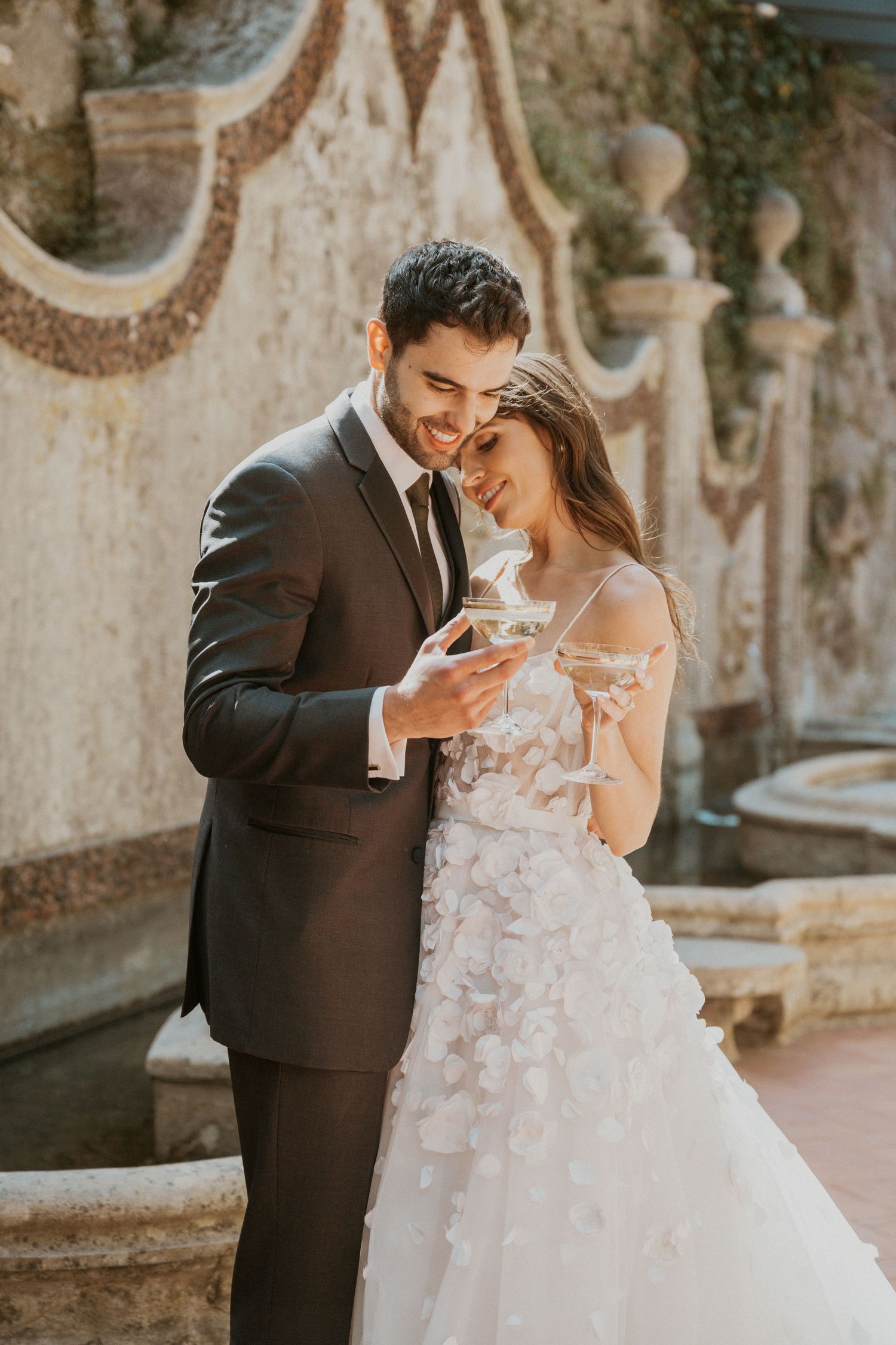 Happy couple in wedding attire sharing champagne glasses outdoors near a stone fountain in warm sunlight.
