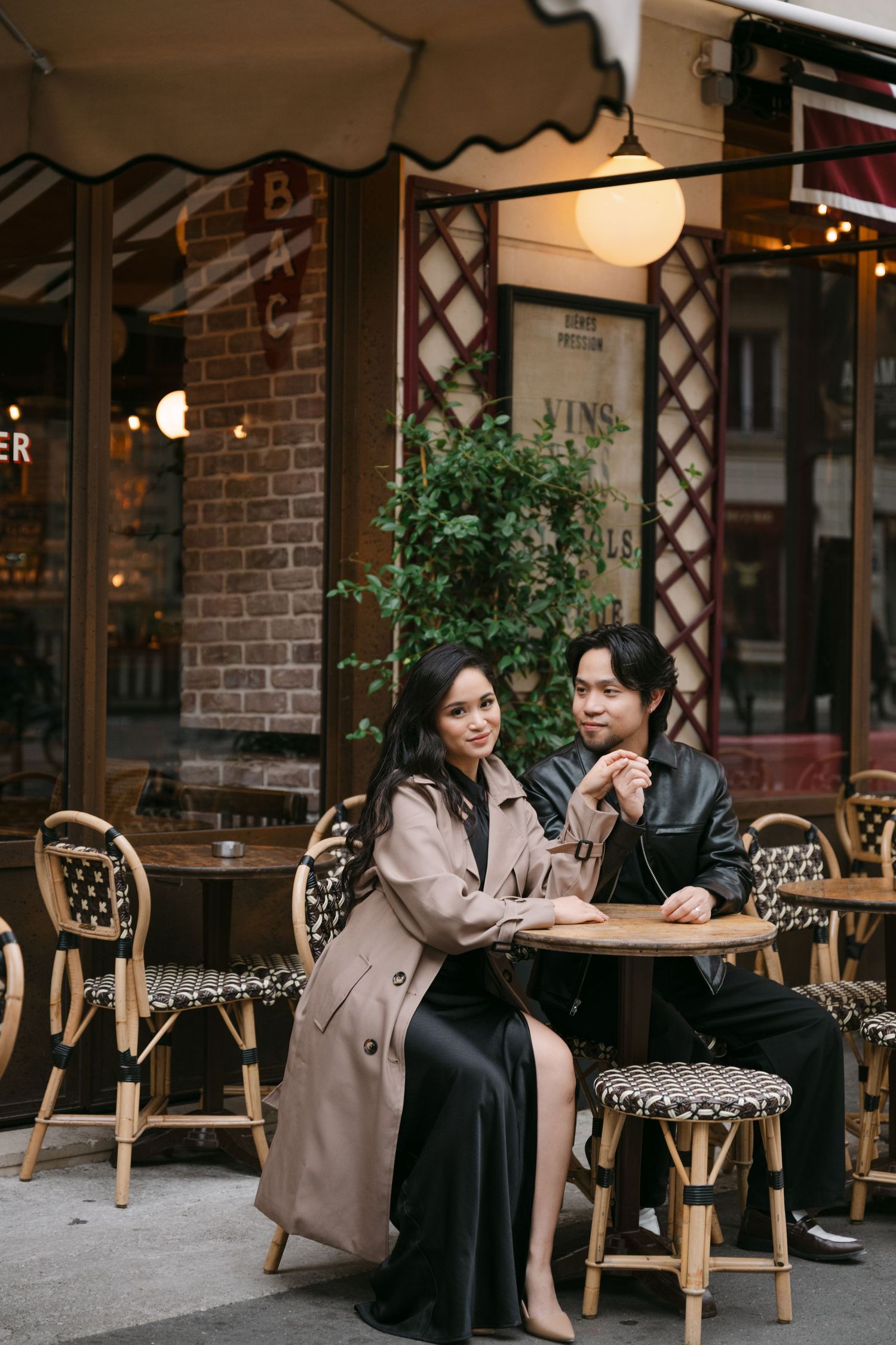 Couple sitting at a Parisian cafe terrace, woman in trench coat and black dress.
