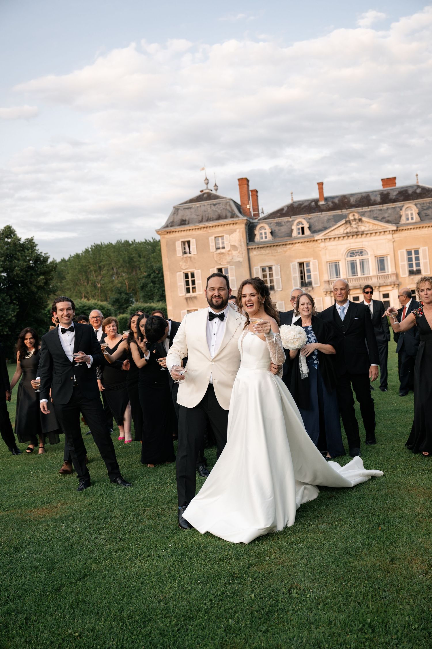 Wedding party celebrates on the lawn of an elegant French chateau with the newlyweds in white at center.
