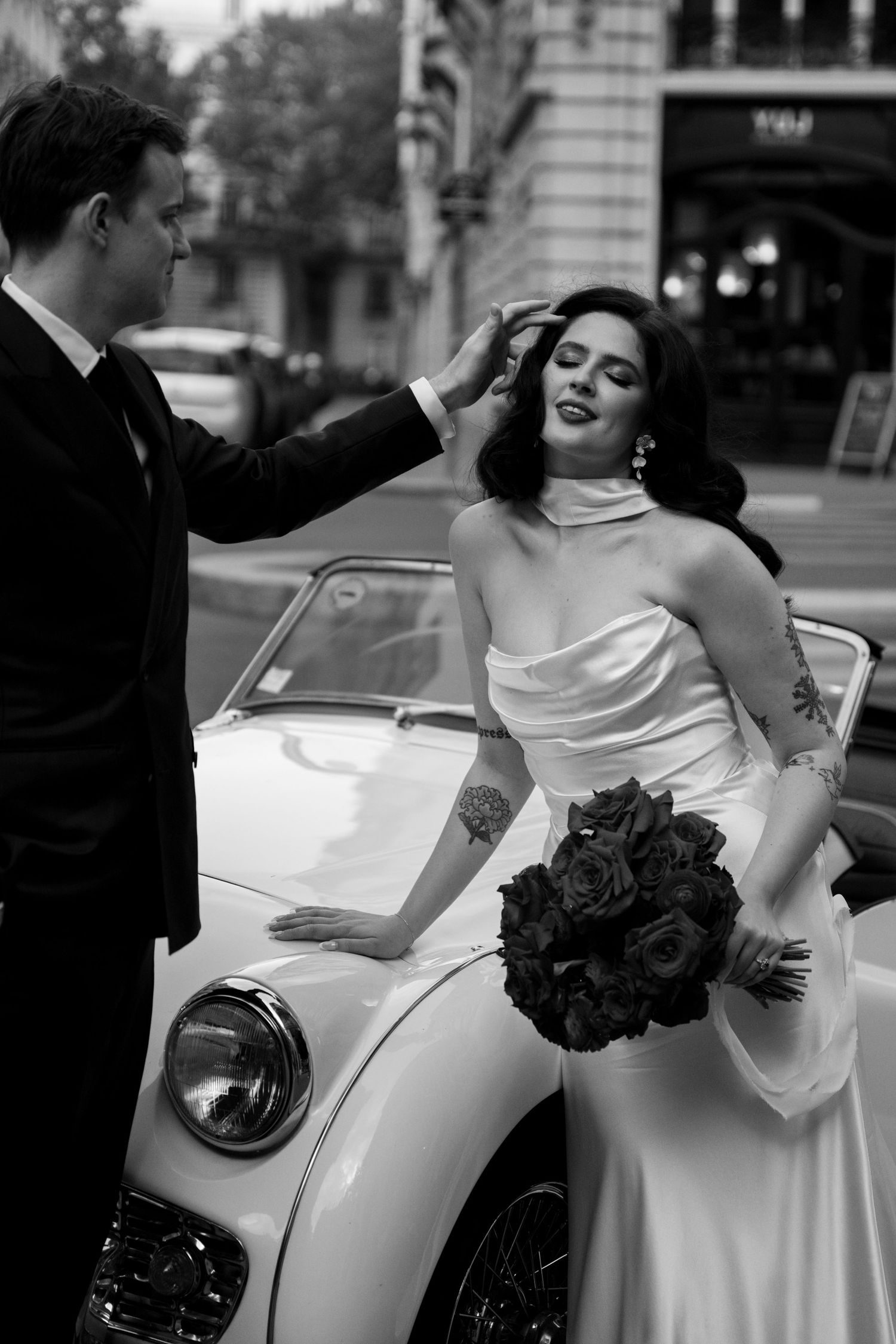Black and white photo of a groom tenderly touching his brides hair beside a vintage white convertible car.