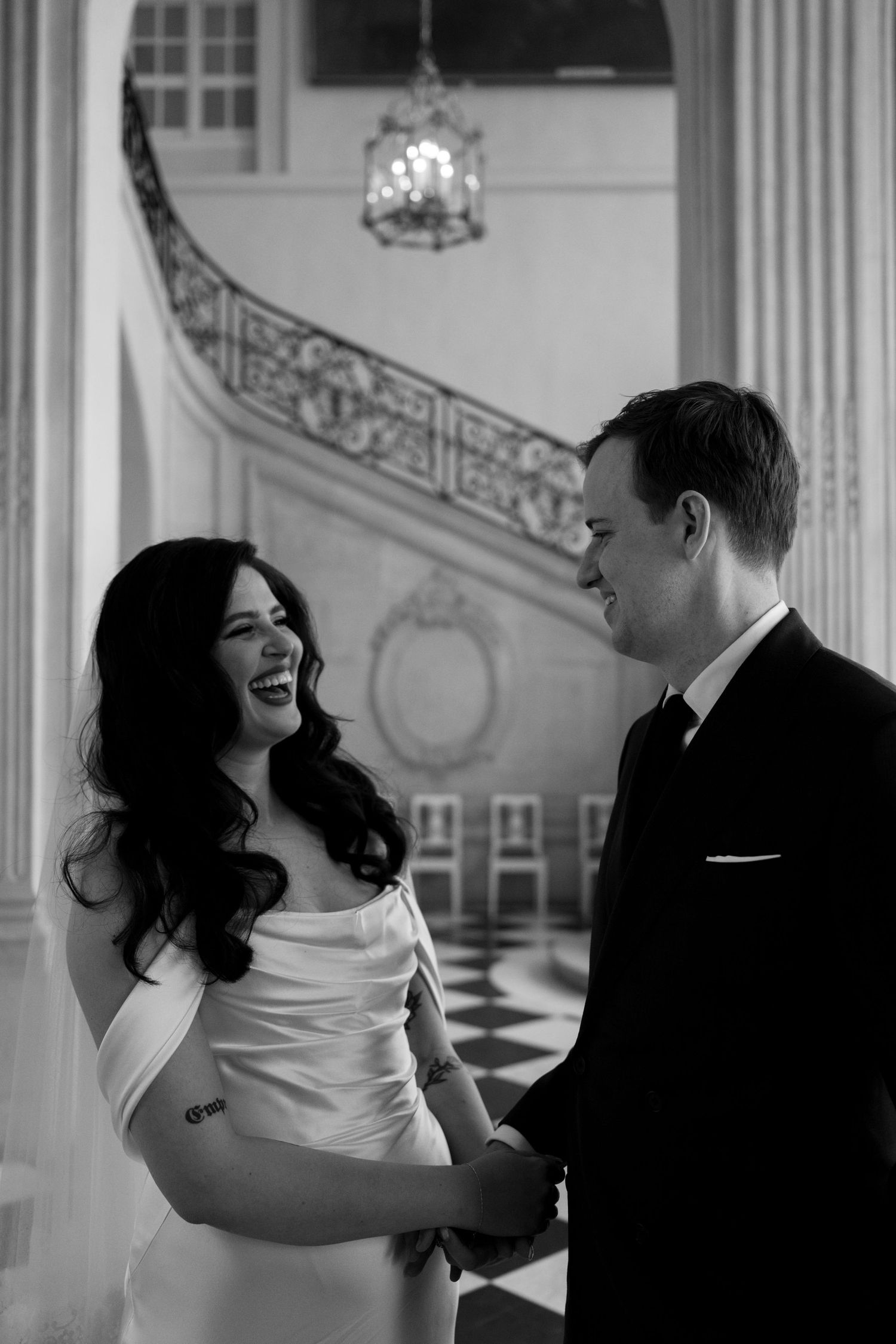 Black and white photo of laughing bride and groom holding hands indoors.