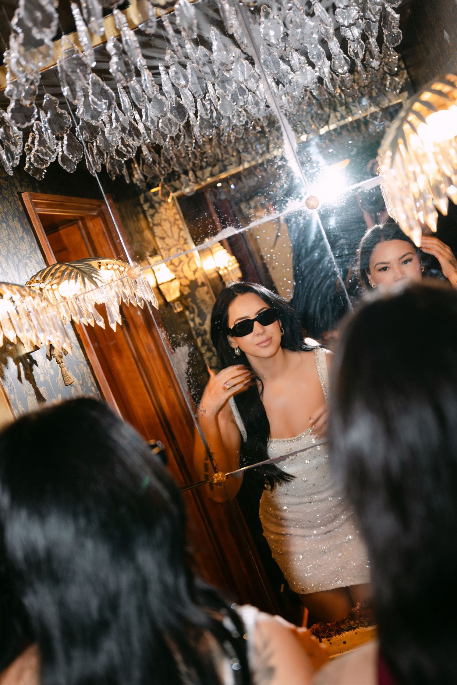 Woman in sparkly dress and sunglasses smiles at mirror reflection in glamorous chandelier-lit venue.