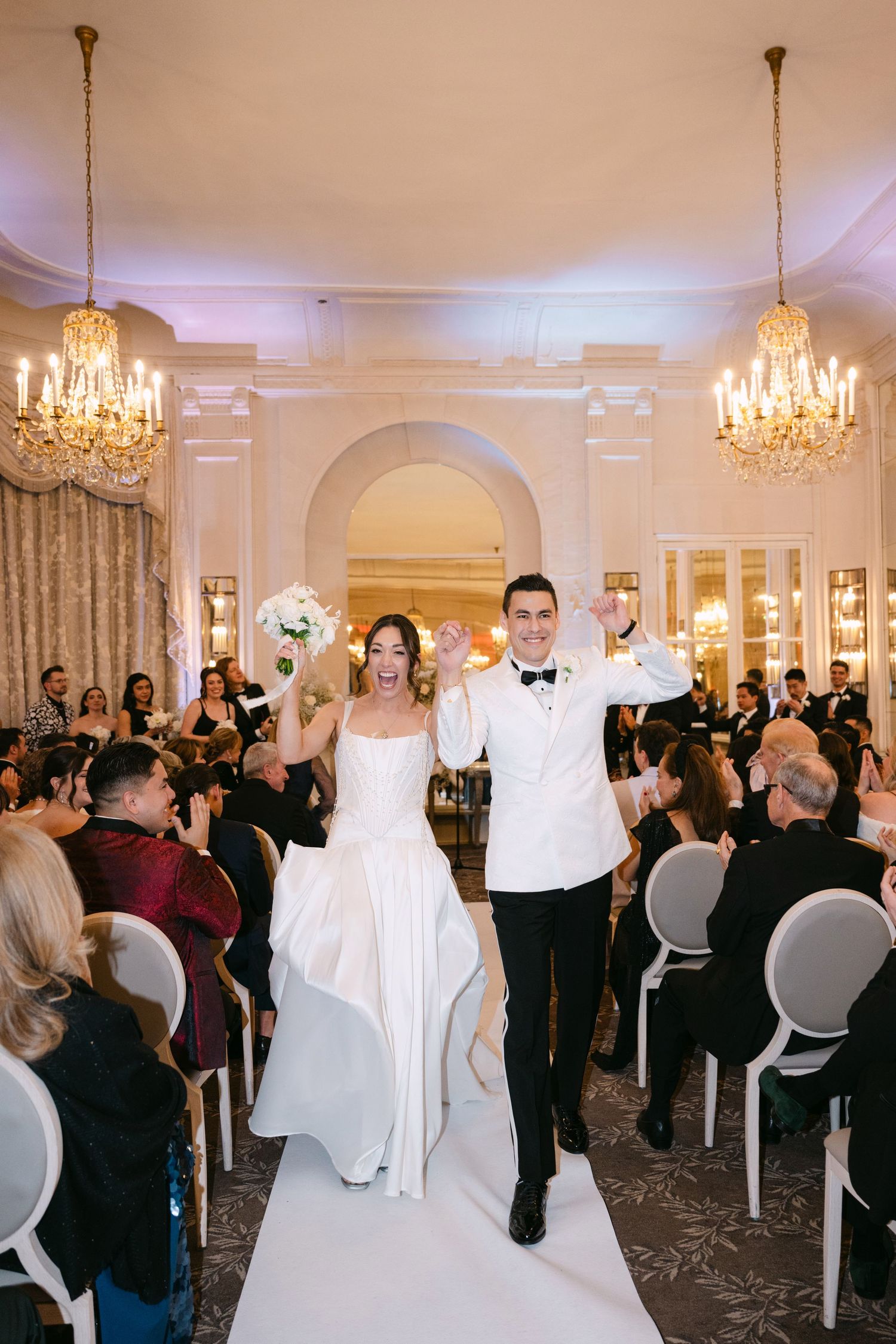 Jubilant newlyweds raise arms while walking down aisle in grand ballroom with crystal chandeliers.