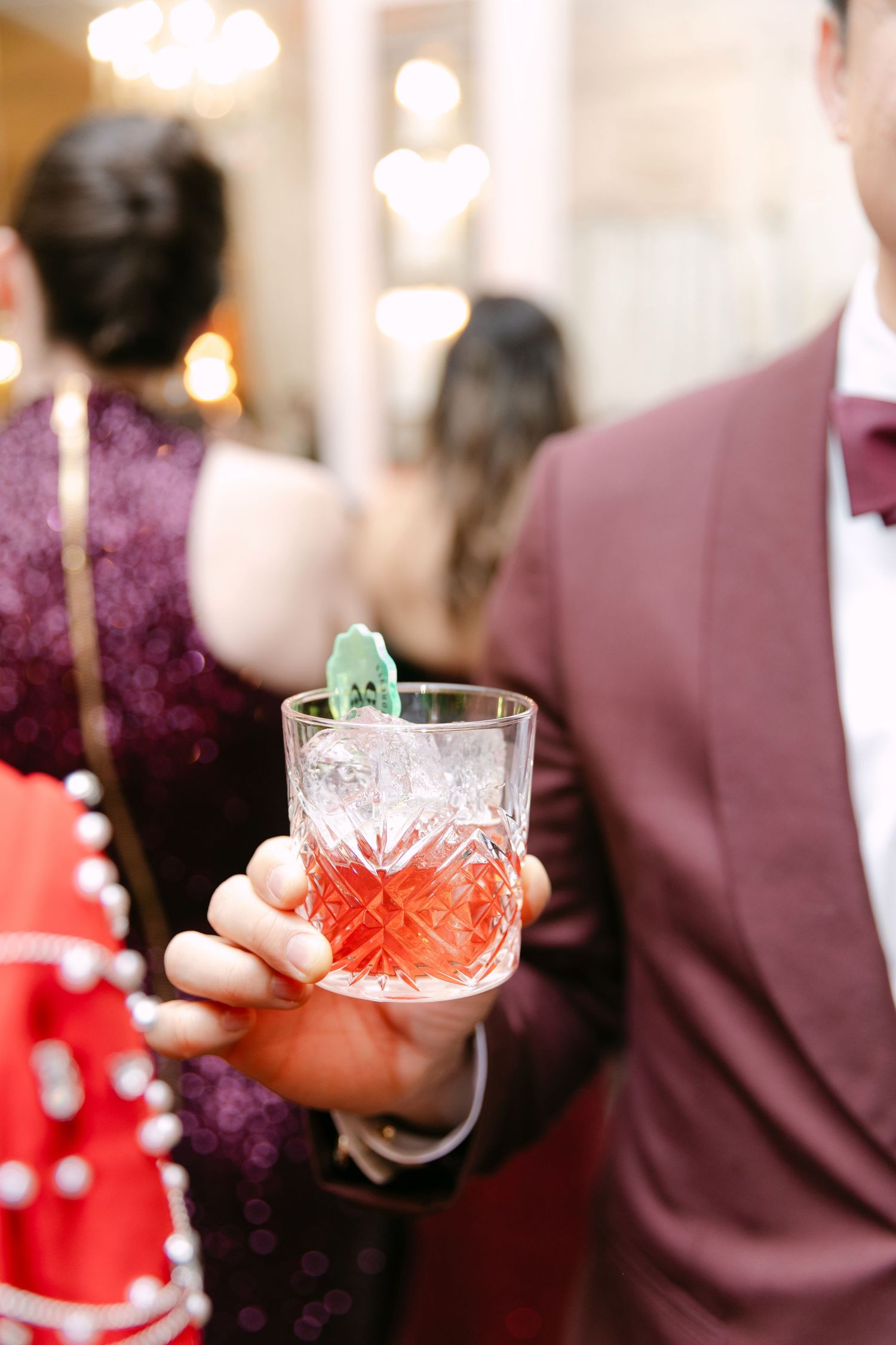 A hand holds a crystal rocks glass with a red cocktail garnished with a small green dinosaur figurine at an elegant event.