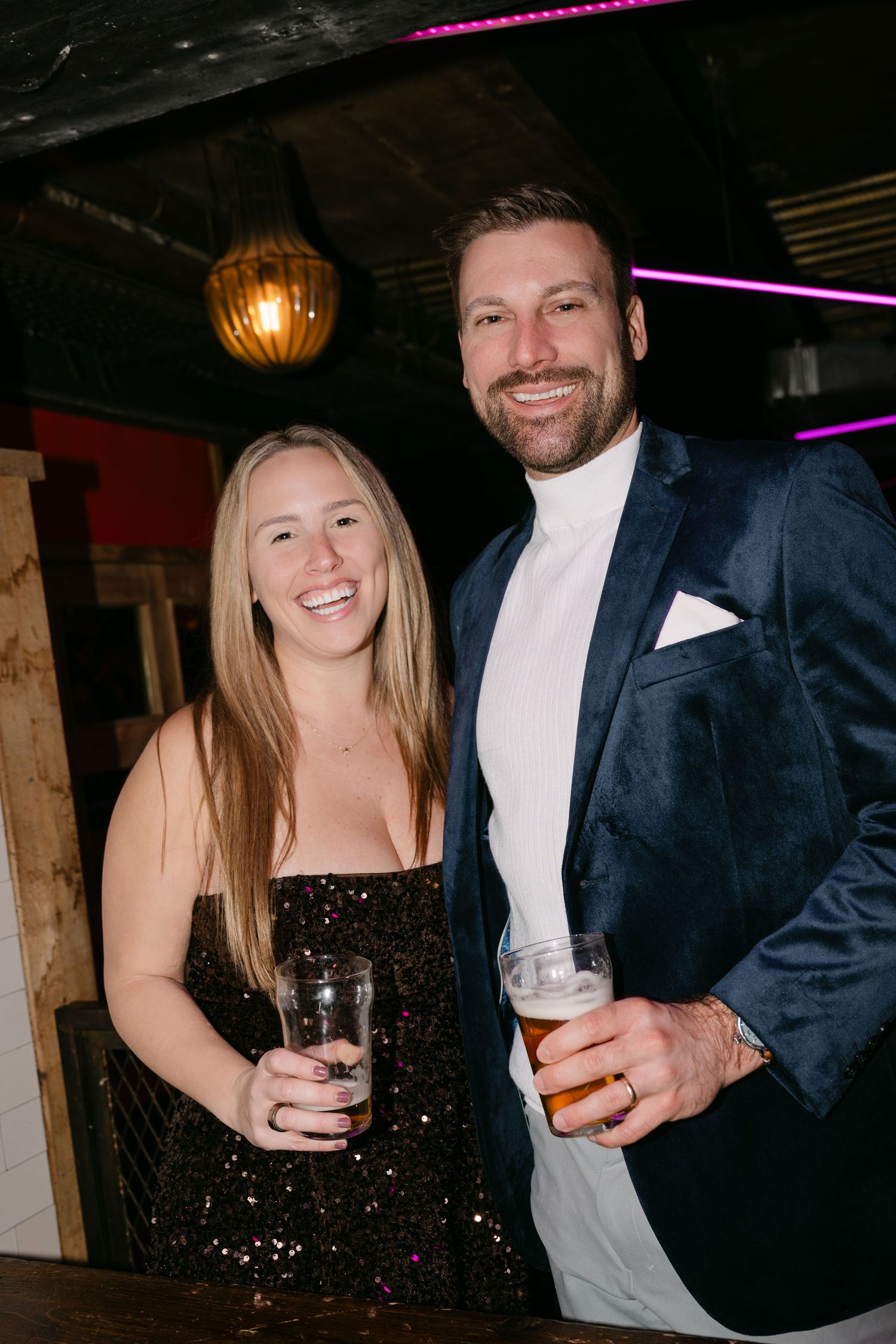 Smiling couple at a party, woman in black sequin dress and man in navy velvet blazer holding drinks.