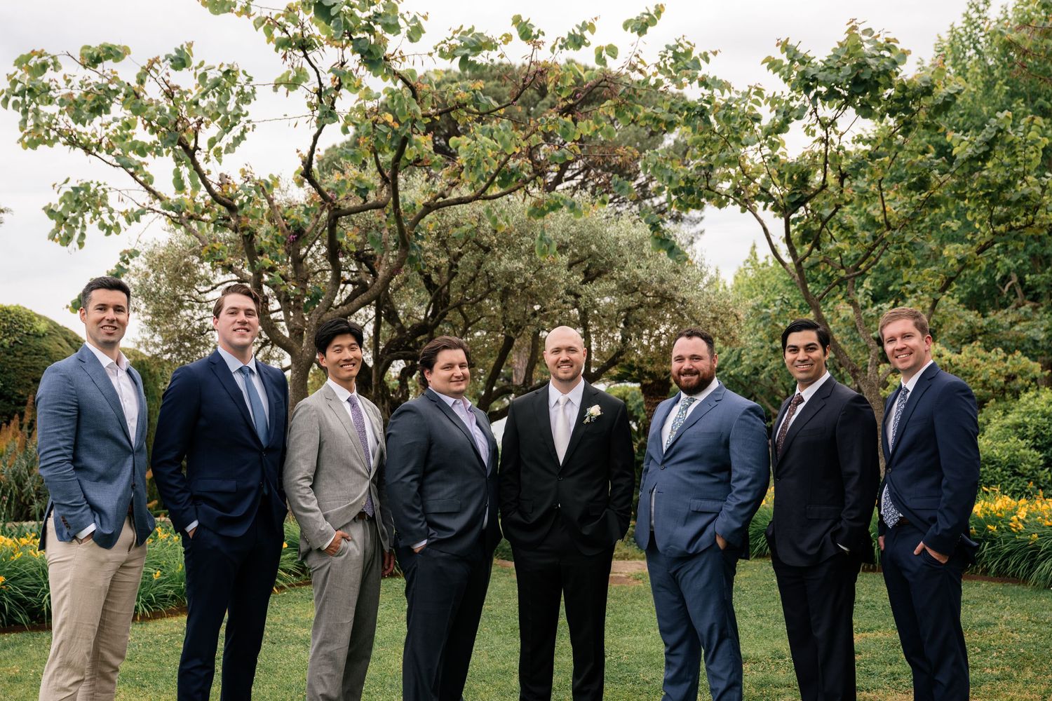 Groom and groomsmen in navy and grey suits pose together in lush green garden for wedding photo.