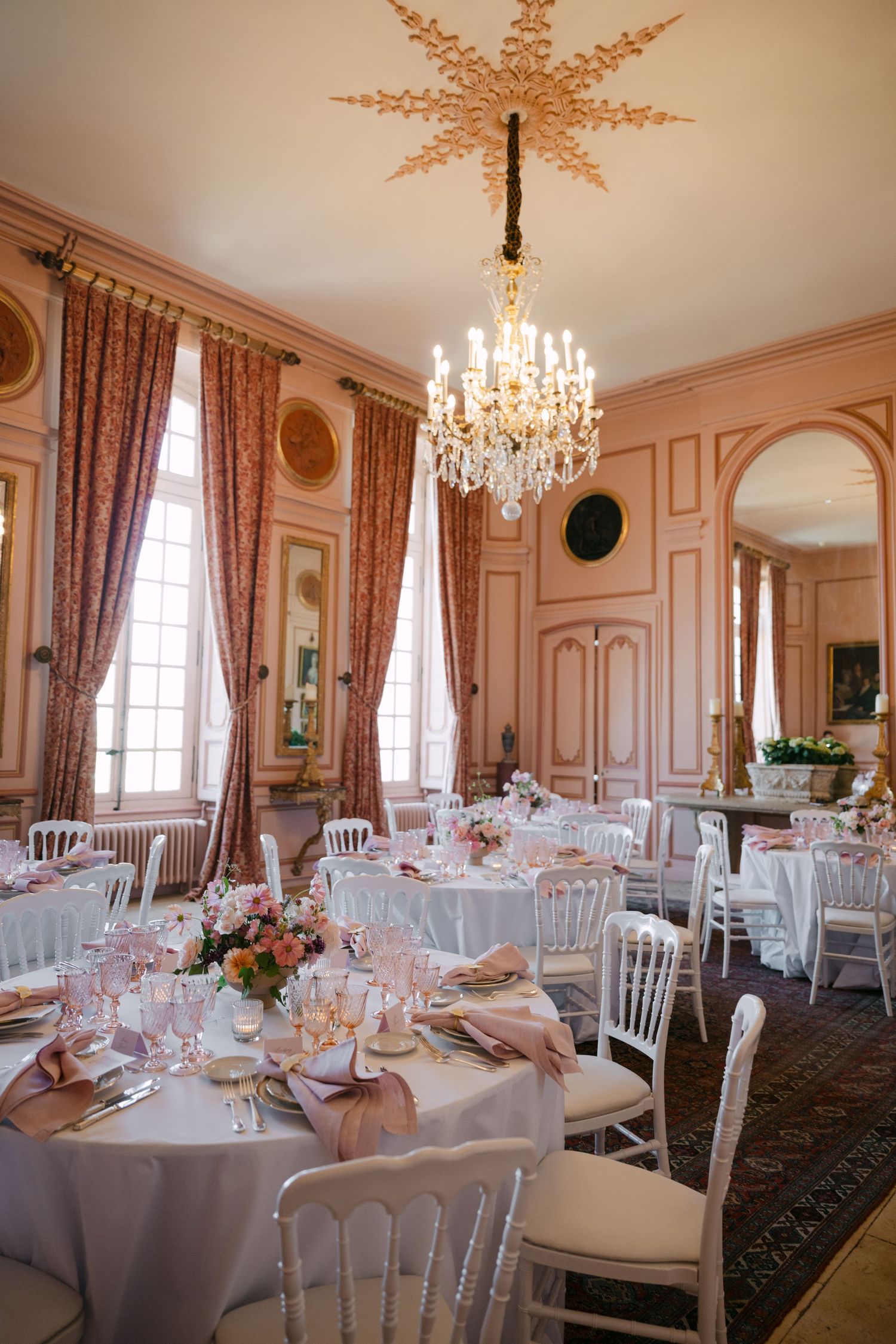 Ornate French chateau dining room with crystal chandelier, pink walls, and elegantly set round tables for a wedding reception.