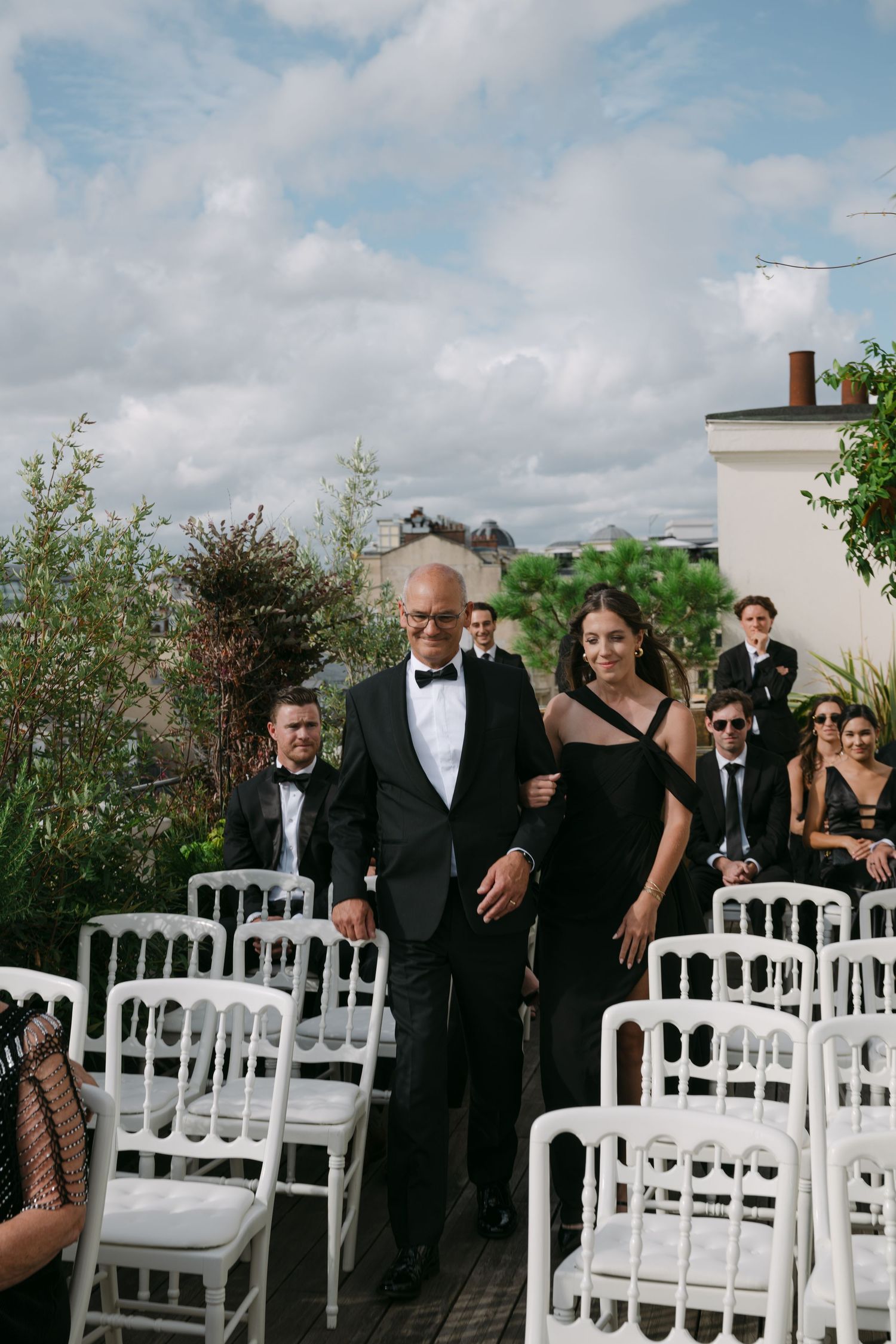 Couple in formal attire walks down outdoor aisle between white chairs at Paris rooftop wedding.